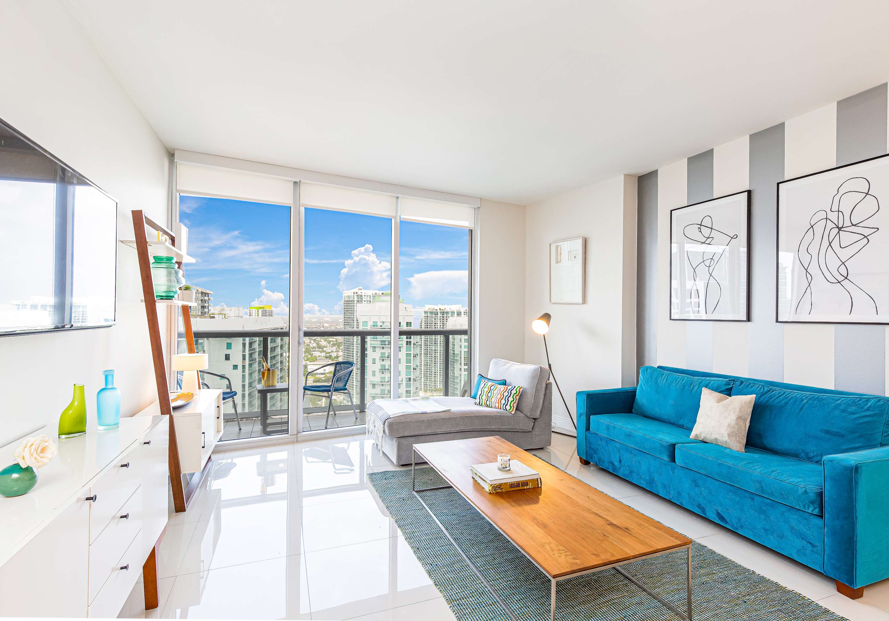 A bright, modern living room with white tiled floors, a vibrant blue sofa, a grey chaise lounge, and a wooden coffee table. Floor-to-ceiling windows offer panoramic city views and a glimpse of a balcony.