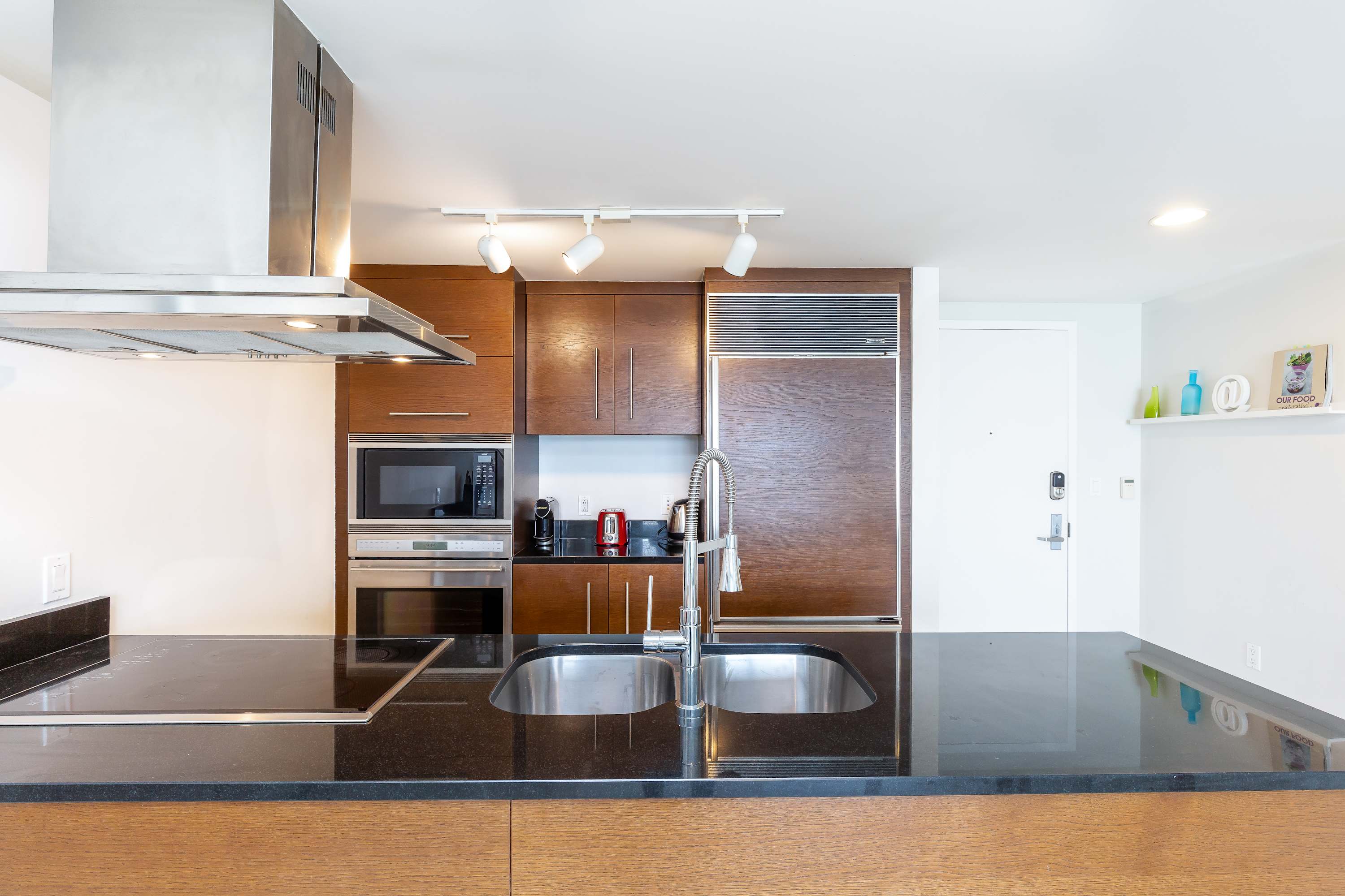 A modern kitchen featuring dark wood cabinetry, black granite countertops, and stainless steel appliances including a microwave, oven, and range hood. A double sink with a pull-down faucet is visible on the island.