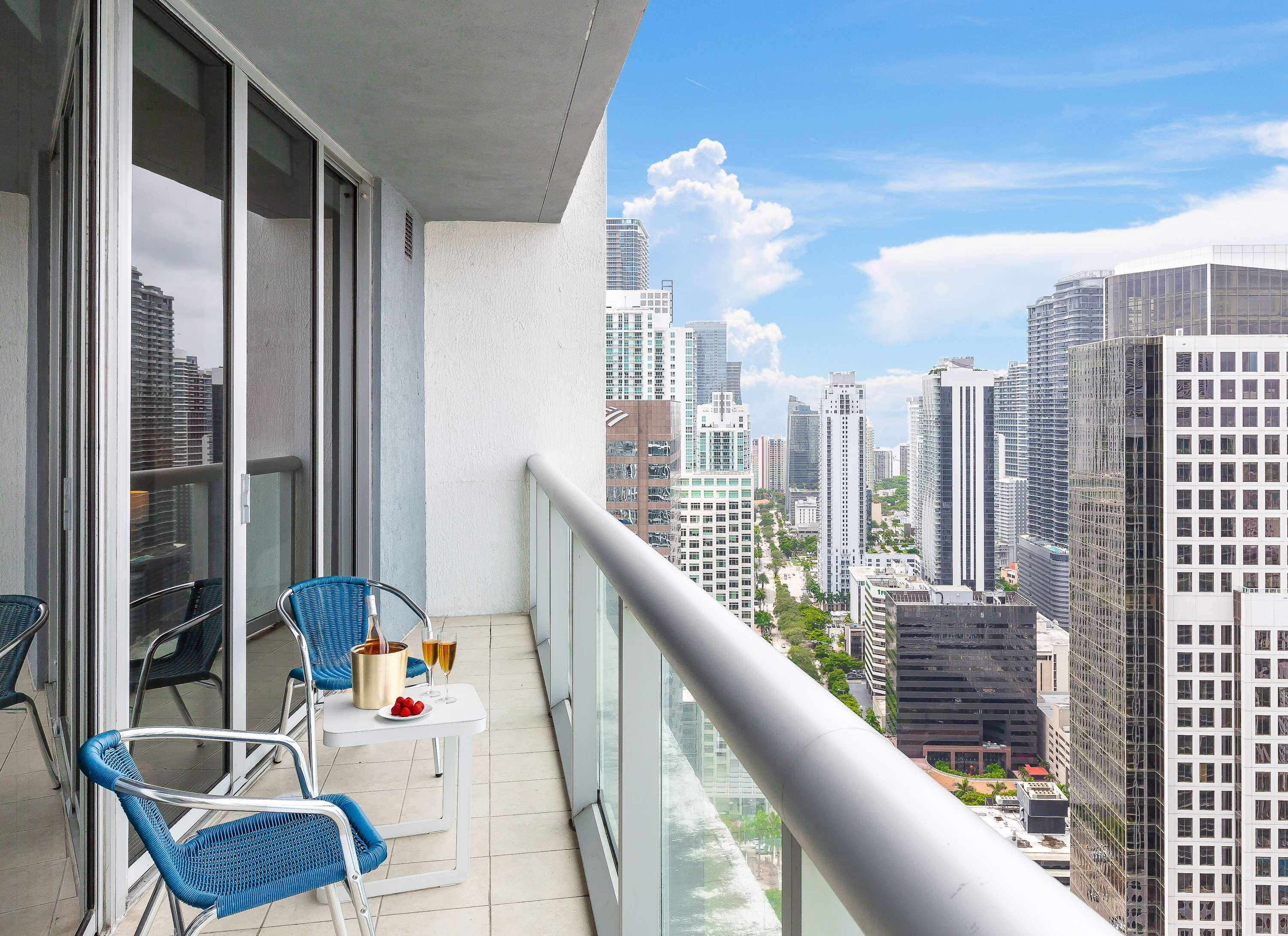 A private balcony with two blue chairs and a small white table, offering expansive views of the city skyline and surrounding high-rise buildings. A bottle of champagne and strawberries are on the table.