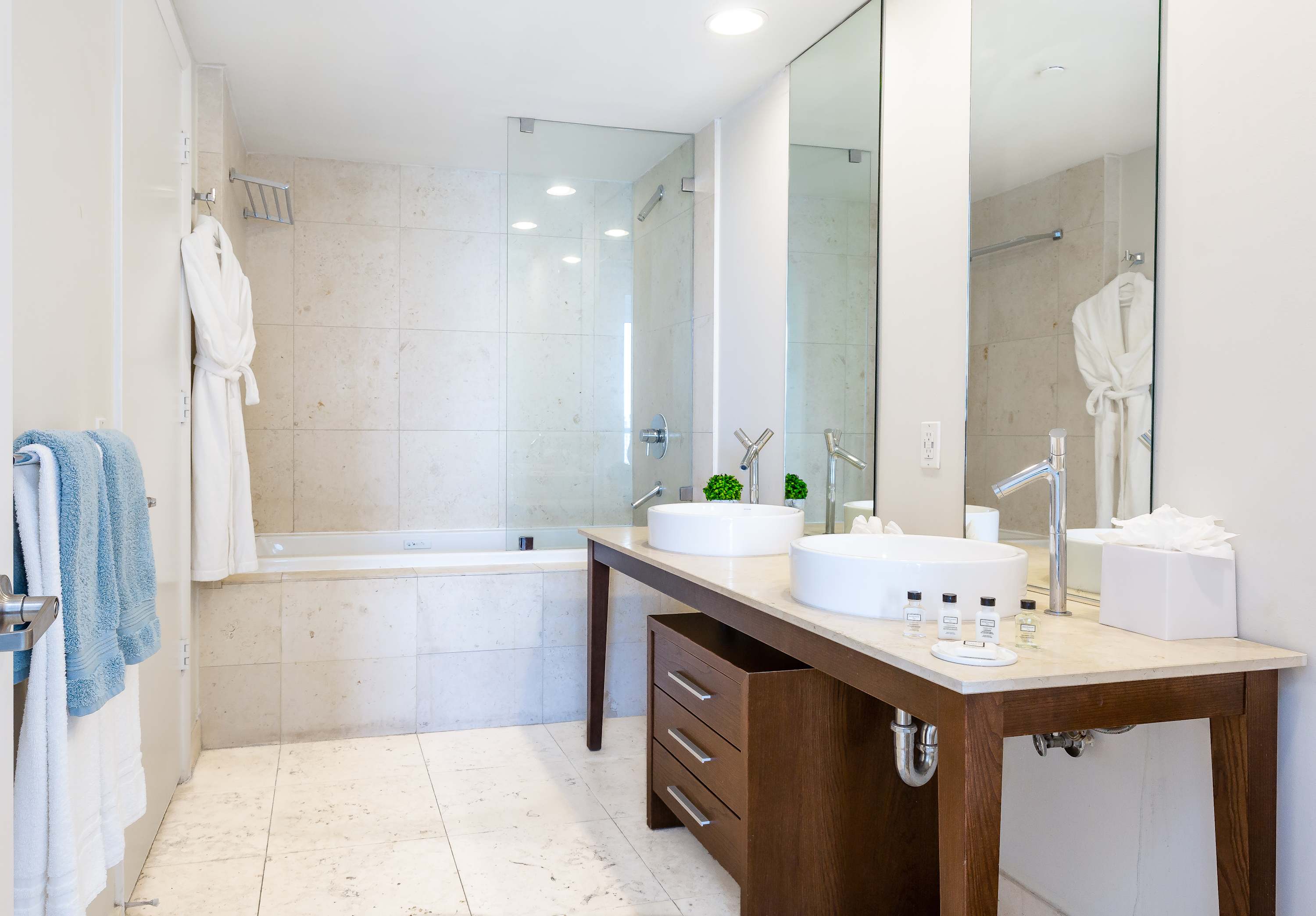 A spacious modern bathroom with light tiled walls and floor, featuring a double vanity with vessel sinks and a large mirror. A bathtub is integrated into a tiled surround, adjacent to a glass-enclosed shower.