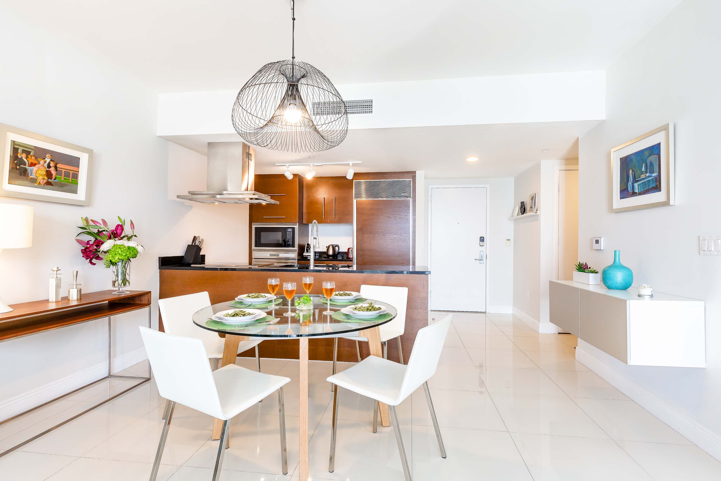 A modern dining area with a round glass table and four white chairs, adjacent to an open-concept kitchen with dark wood cabinetry and stainless steel appliances. A unique wire chandelier hangs above the table.