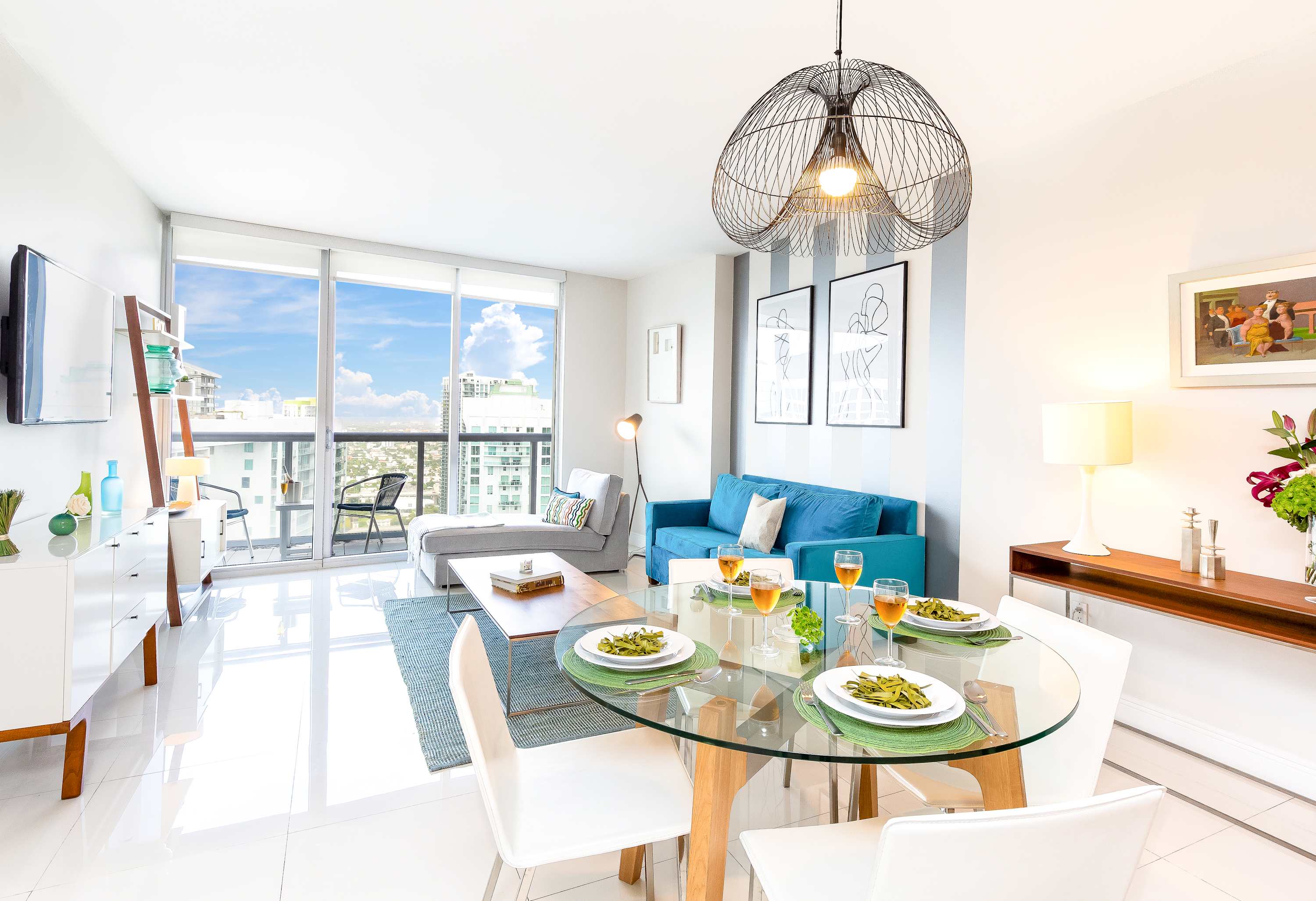 An open-concept living and dining area with white tiled floors, featuring a glass-top dining table with white chairs and a unique wire pendant light. The space includes a blue sofa and floor-to-ceiling windows with city views.