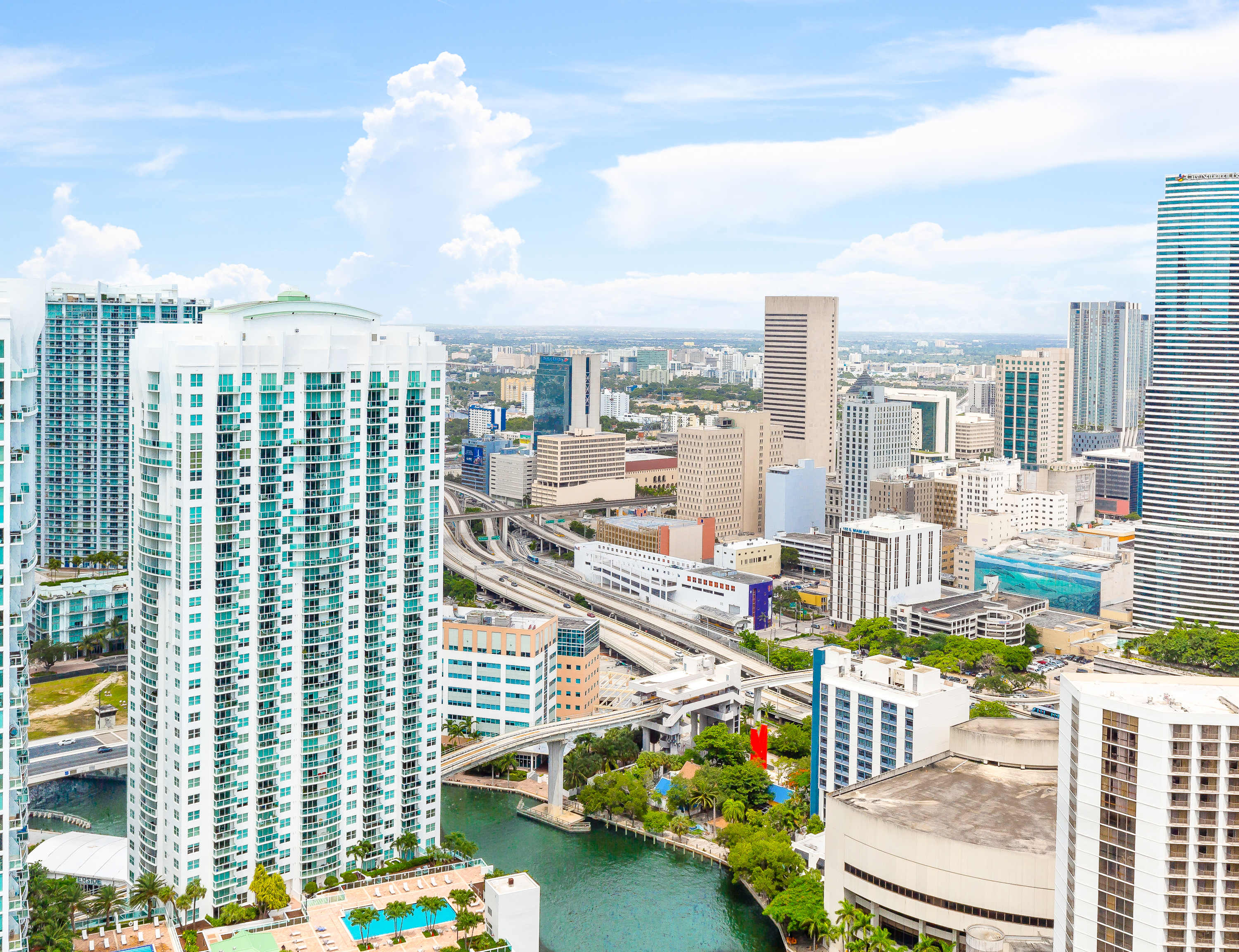 An aerial view of a dense urban cityscape with numerous high-rise buildings, a river, and elevated highways under a partly cloudy sky. The image highlights the scale and density of the city.