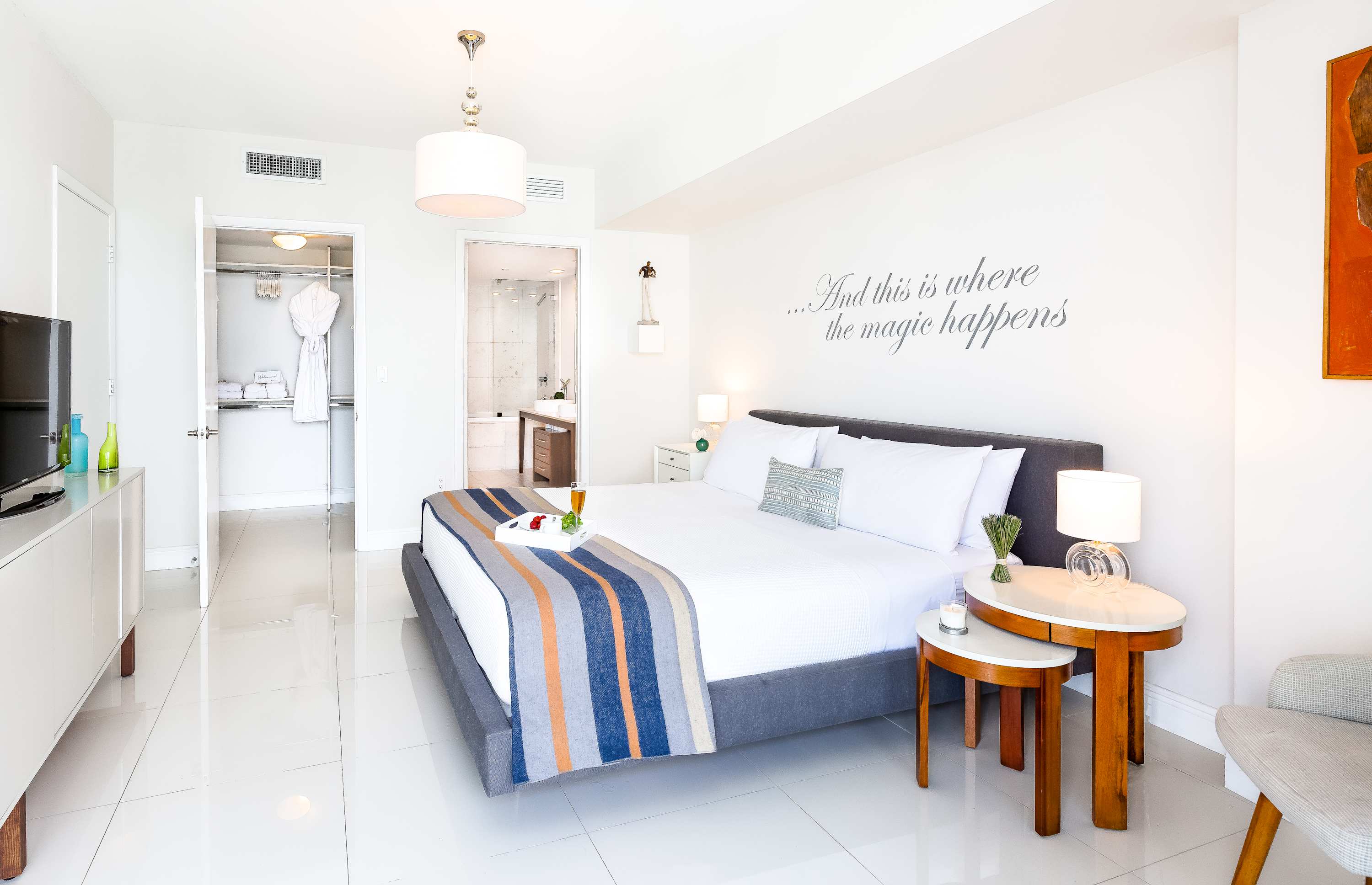 A spacious bedroom with a king bed, white tiled floors, and a walk-in closet visible through an open door. The room is decorated with a wall decal and modern lighting fixtures.
