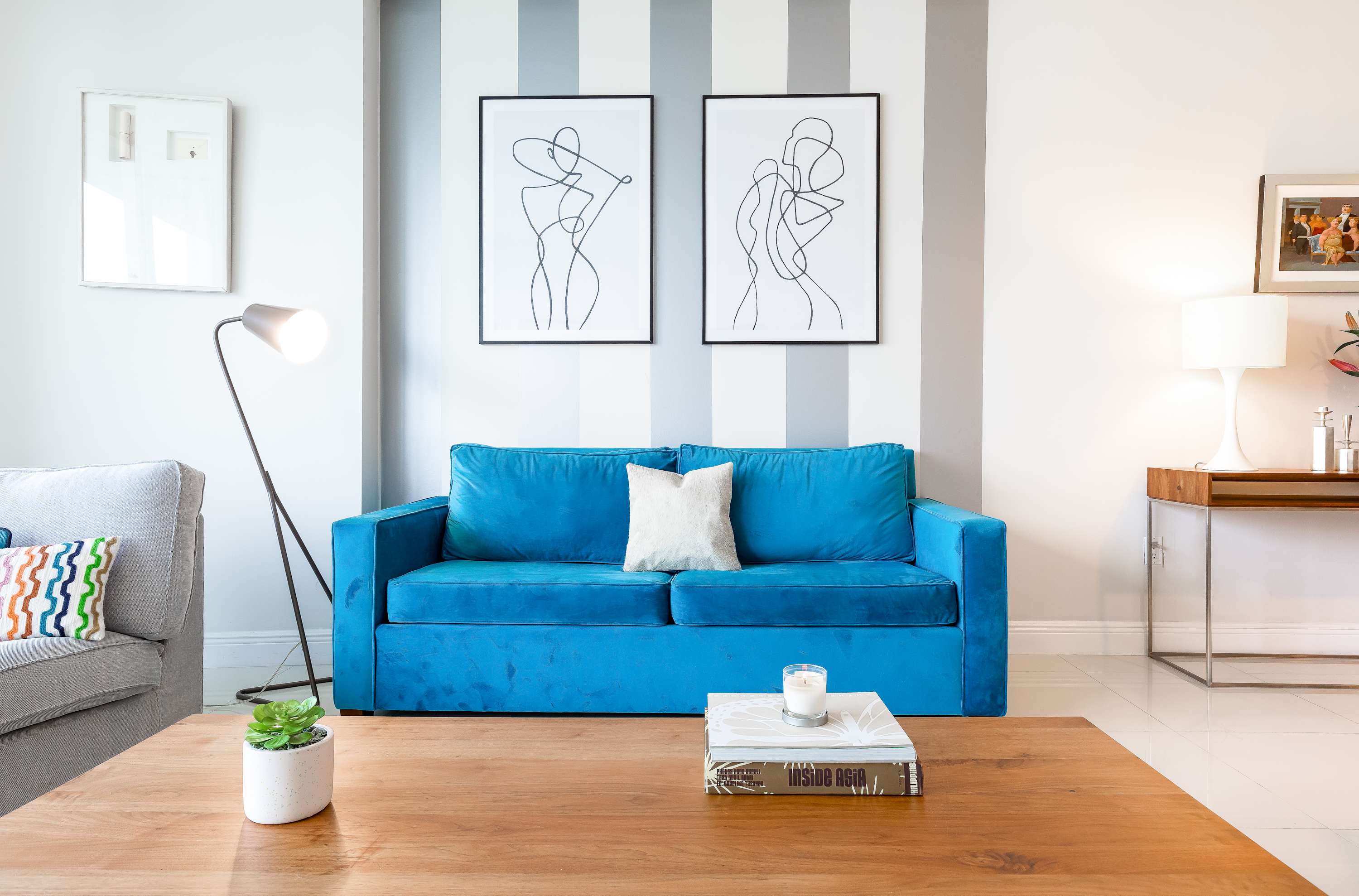 A cozy living area featuring a vibrant teal velvet sofa, a light wood coffee table, and a grey armchair, set against a white wall with grey and white stripes and framed abstract art. A modern floor lamp provides ambient lighting.