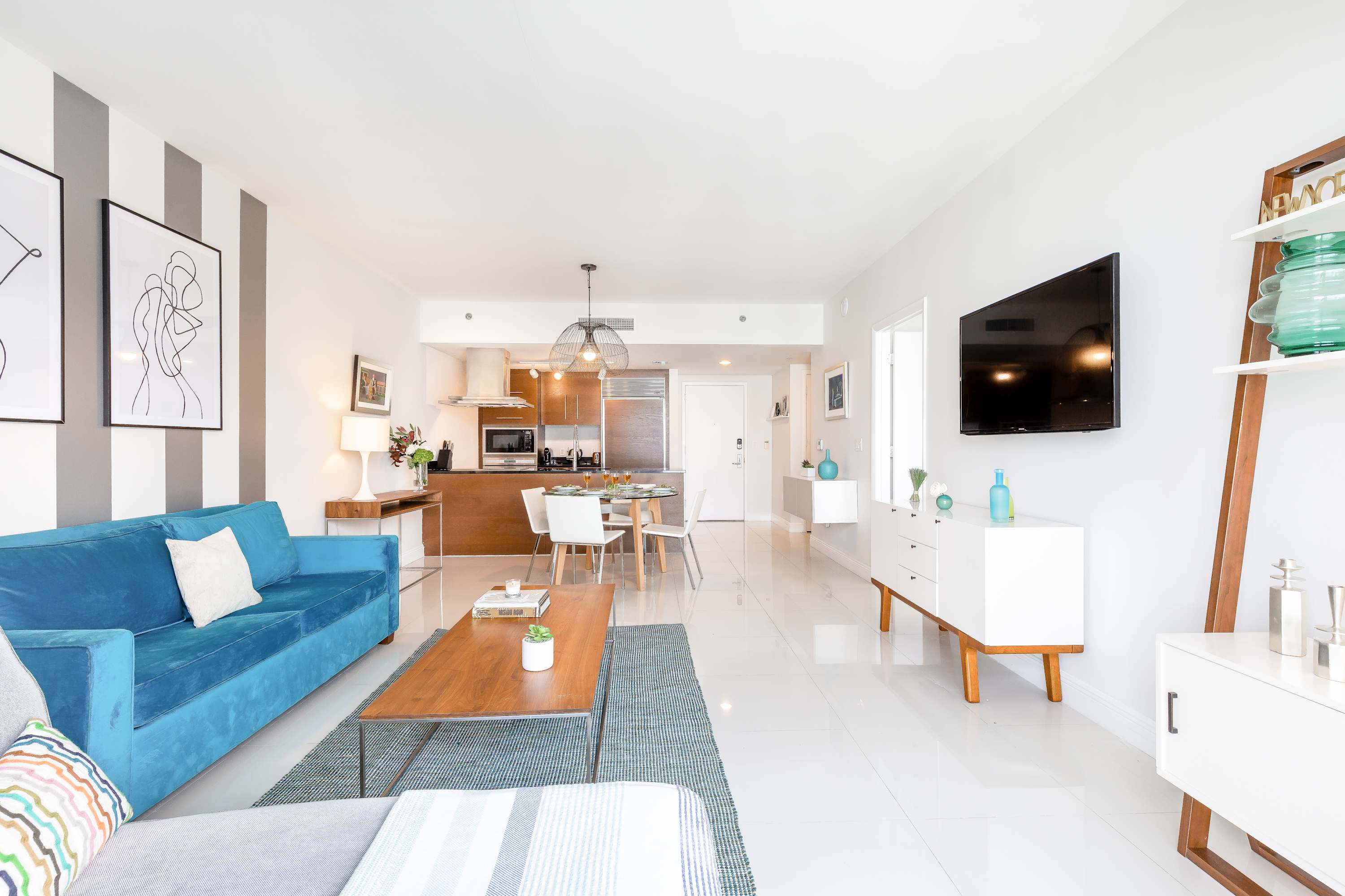 An open-concept living space with white tiled floors, a teal sofa, a wooden coffee table, and a white console with a TV. The kitchen and dining area are visible in the background, featuring dark wood cabinetry and a glass dining table.