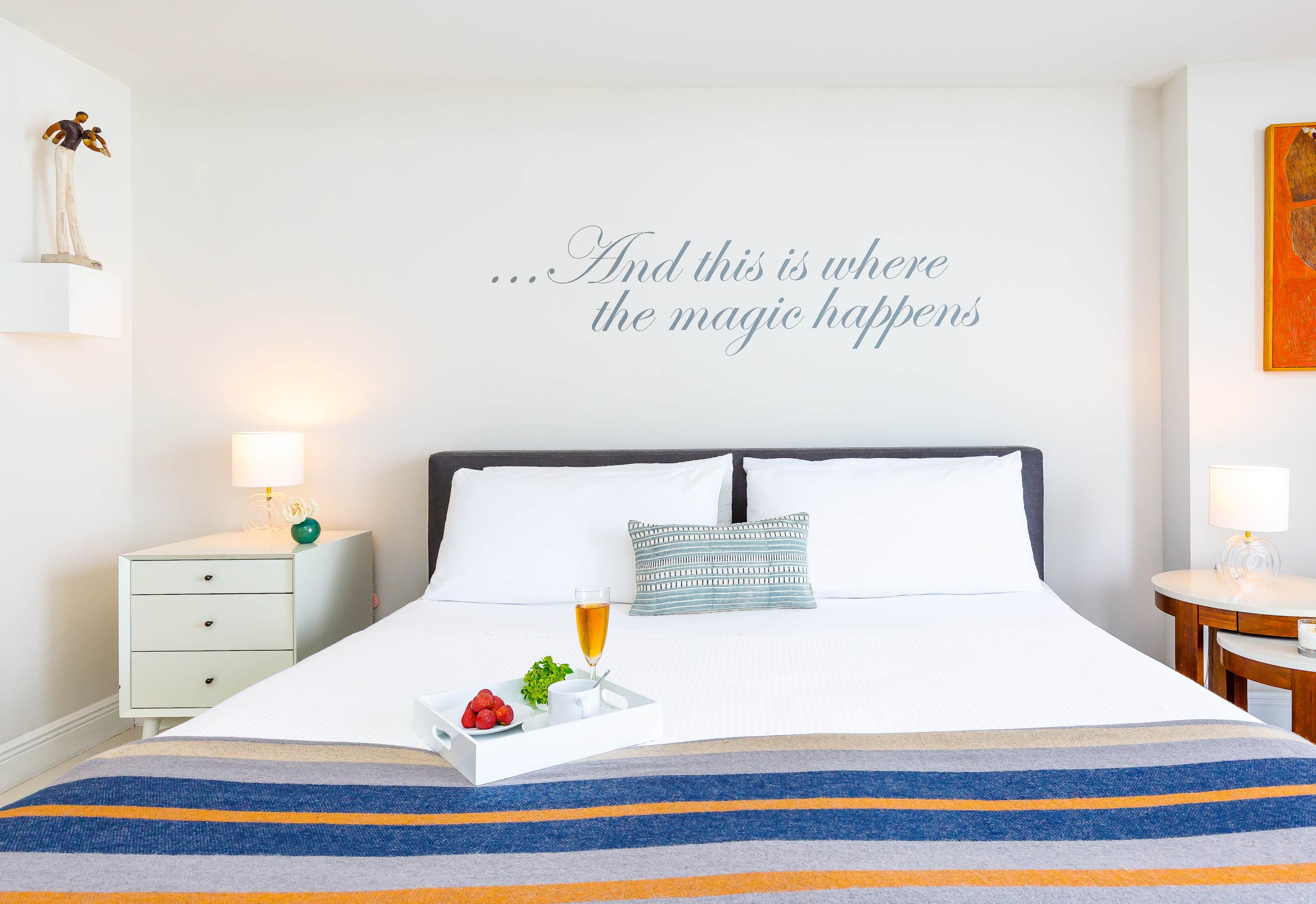 A bright bedroom with a king-sized bed featuring a striped blanket, white tiled floors, and a white nightstand. A wall decal reads "...And this is where the magic happens" above the bed.