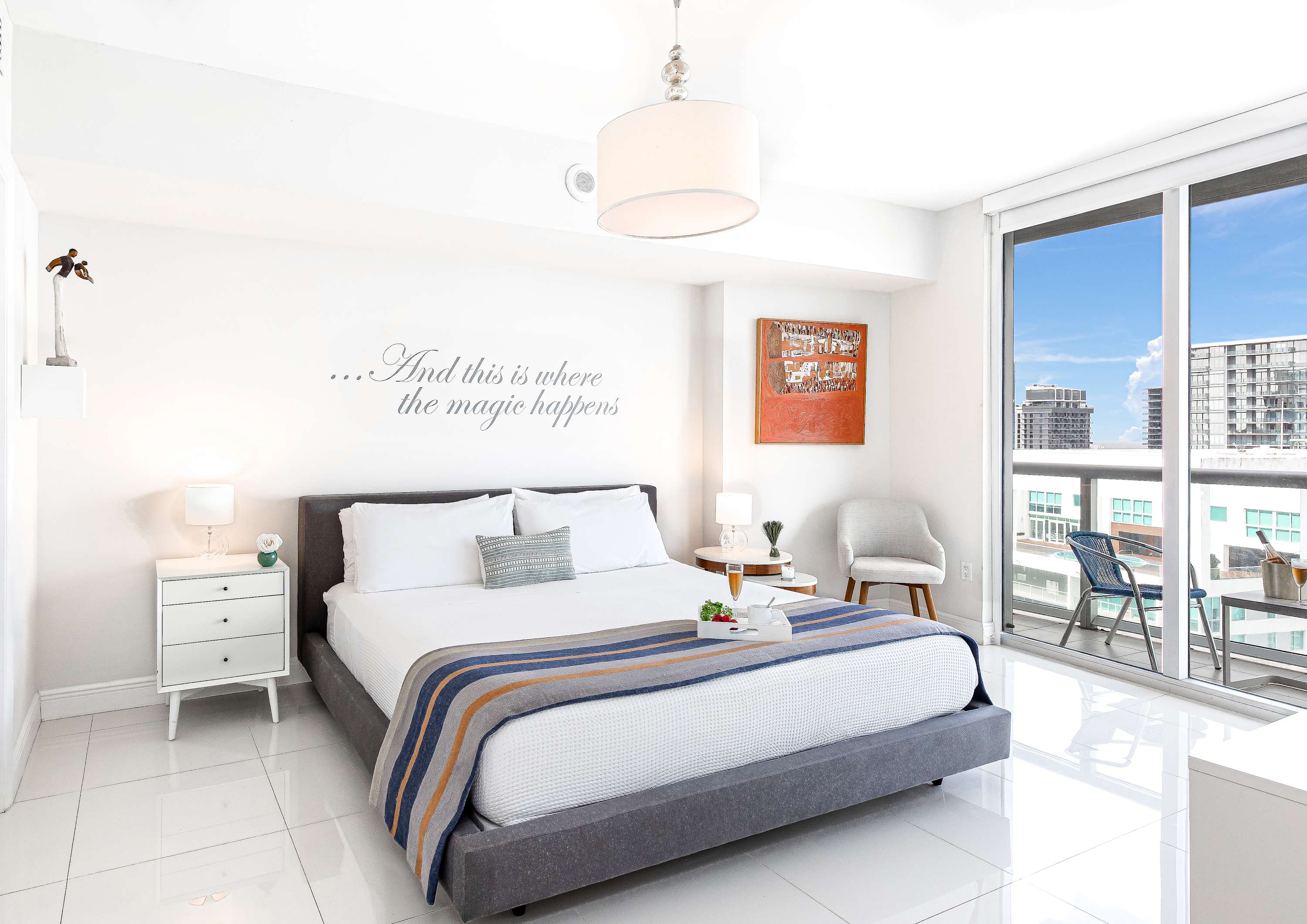 A bright bedroom with a king-sized bed, white tiled floors, and a private balcony offering city views through floor-to-ceiling glass doors. A wall decal with text is visible above the bed.