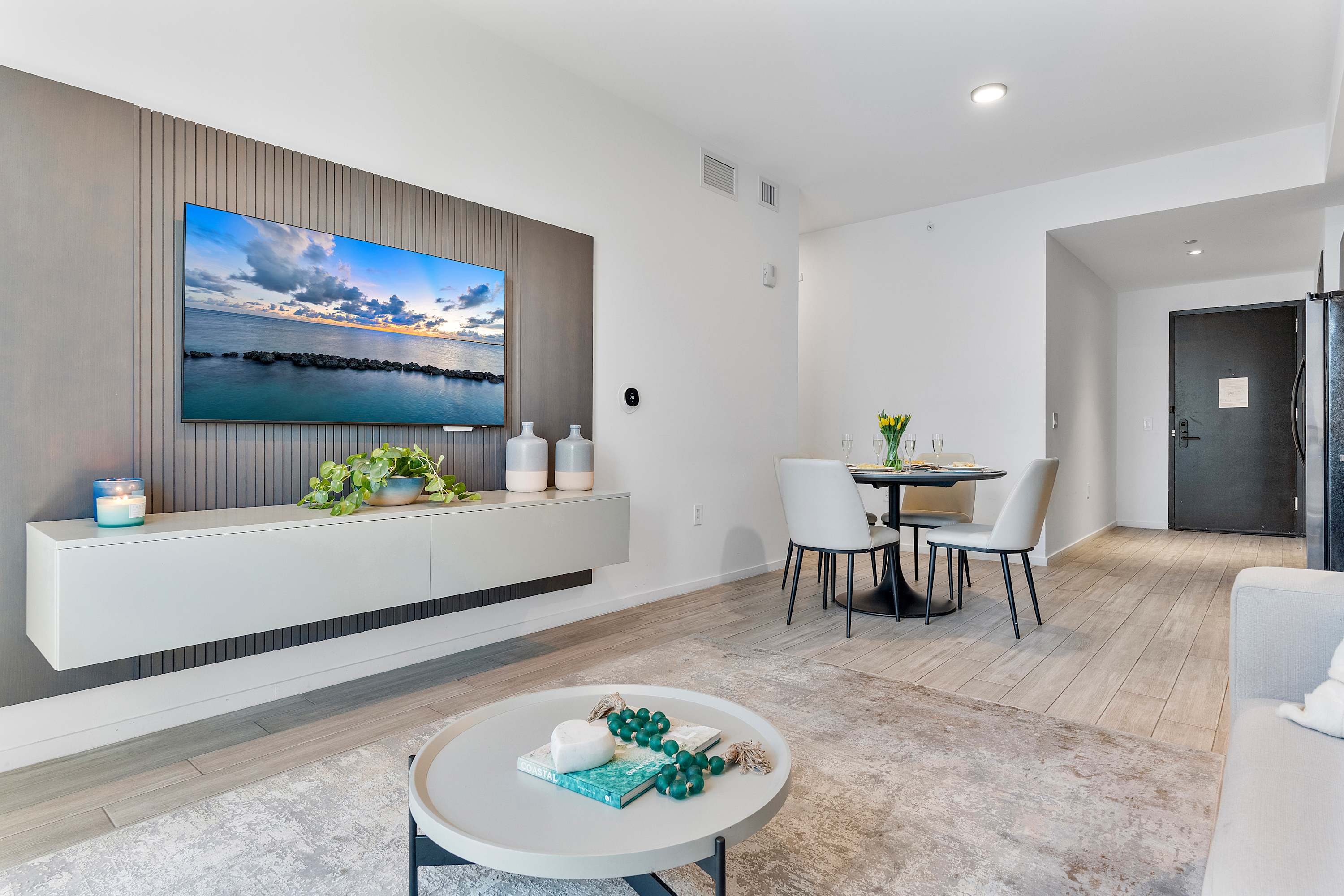 Contemporary living space with a wall-mounted TV above a floating console, a round coffee table, and a dining area with a dark round table and light gray chairs, all on light wood-look tile flooring.