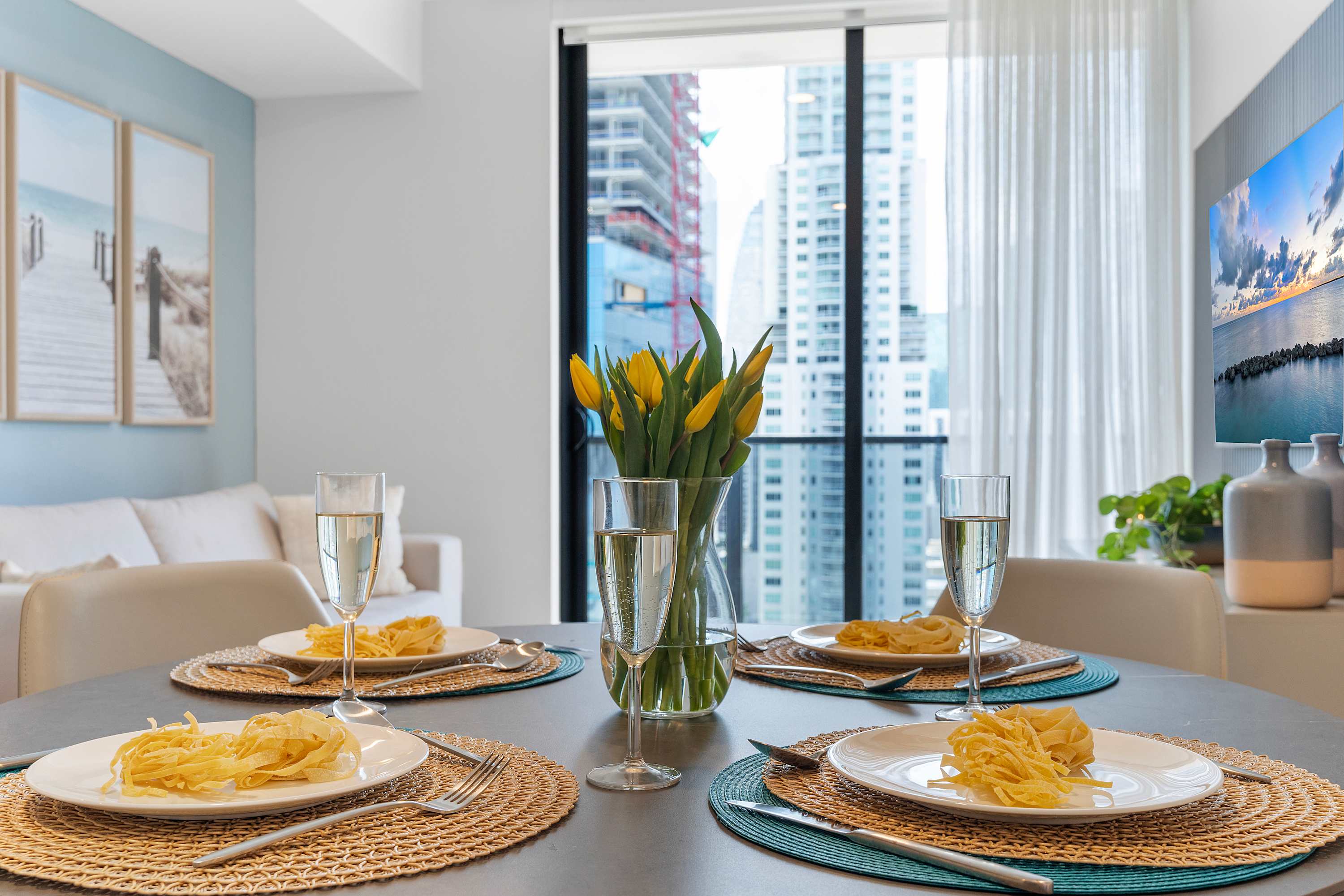 Round dining table set for four with white plates, golden placemats, and champagne flutes, featuring a vase of yellow tulips and a view of city buildings through large windows.