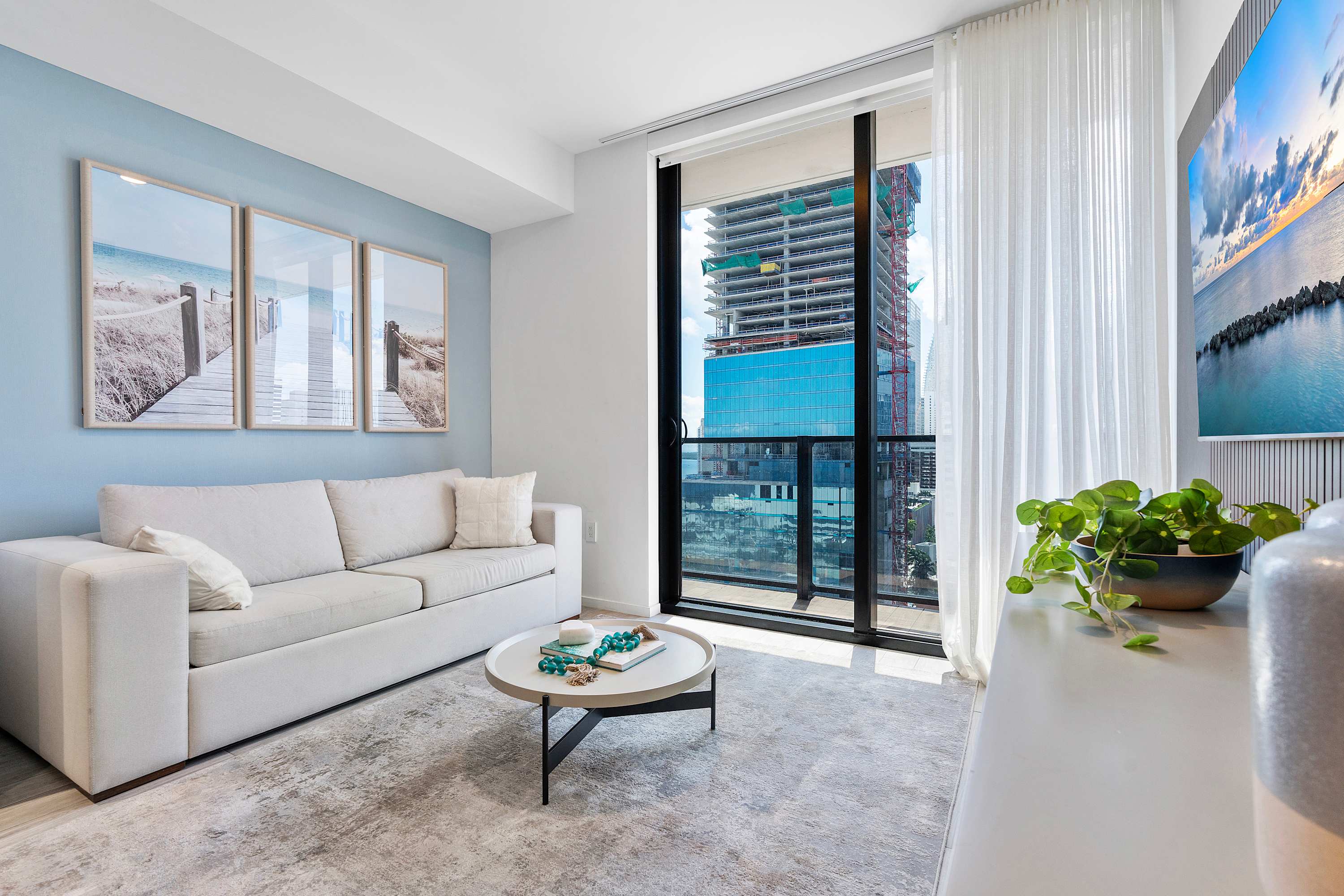 Bright living room with a light-colored sofa, a round coffee table, and a light blue accent wall adorned with three framed beach-themed artworks. Large floor-to-ceiling windows offer a view of a city building under construction.