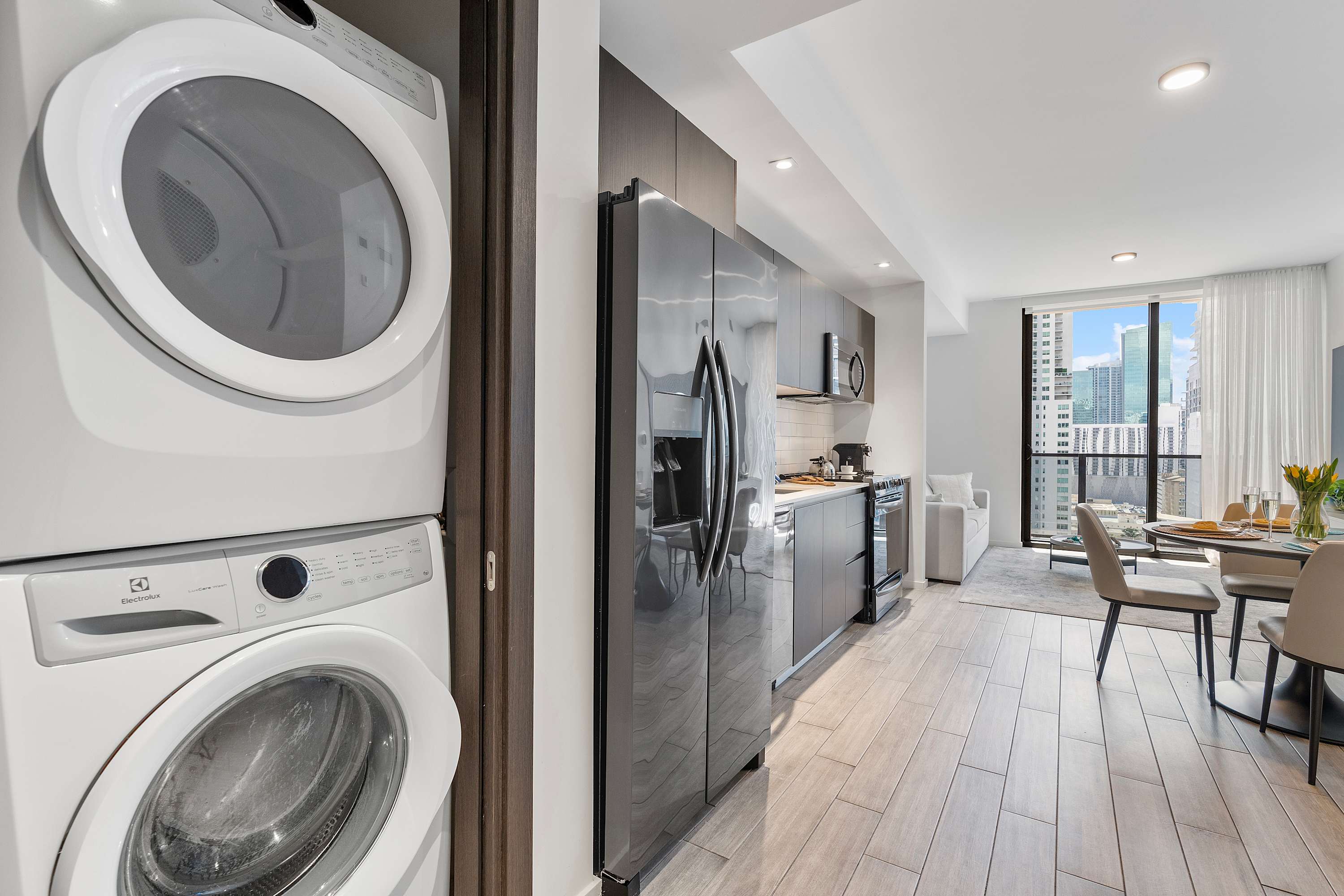 Compact laundry closet with stacked white washer and dryer units, adjacent to a modern kitchen with dark cabinetry and a stainless steel refrigerator, leading to an open living area.