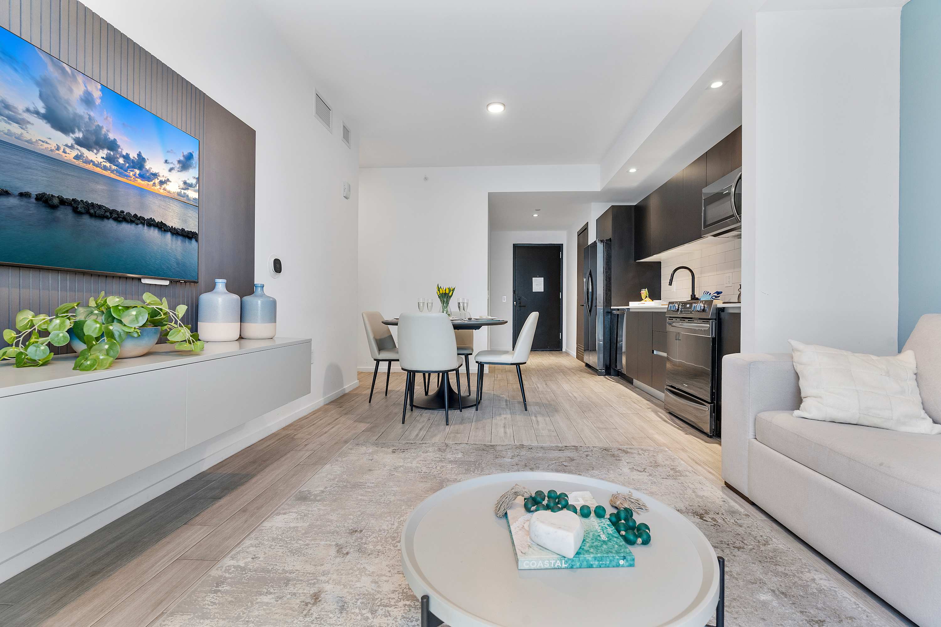 Open-plan living area featuring a light gray sofa, a round coffee table, a dining set, and a modern kitchen with dark cabinetry, all illuminated by natural light from large windows.
