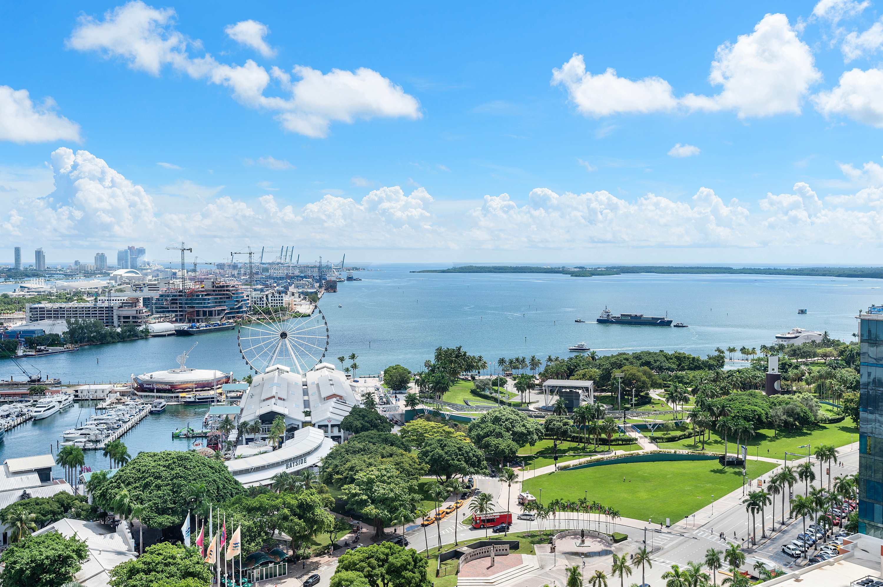 An expansive aerial view captures a vibrant city skyline with numerous high-rise buildings, a large bay dotted with boats, and a prominent Ferris wheel on the waterfront, all under a bright blue sky with fluffy white clouds.