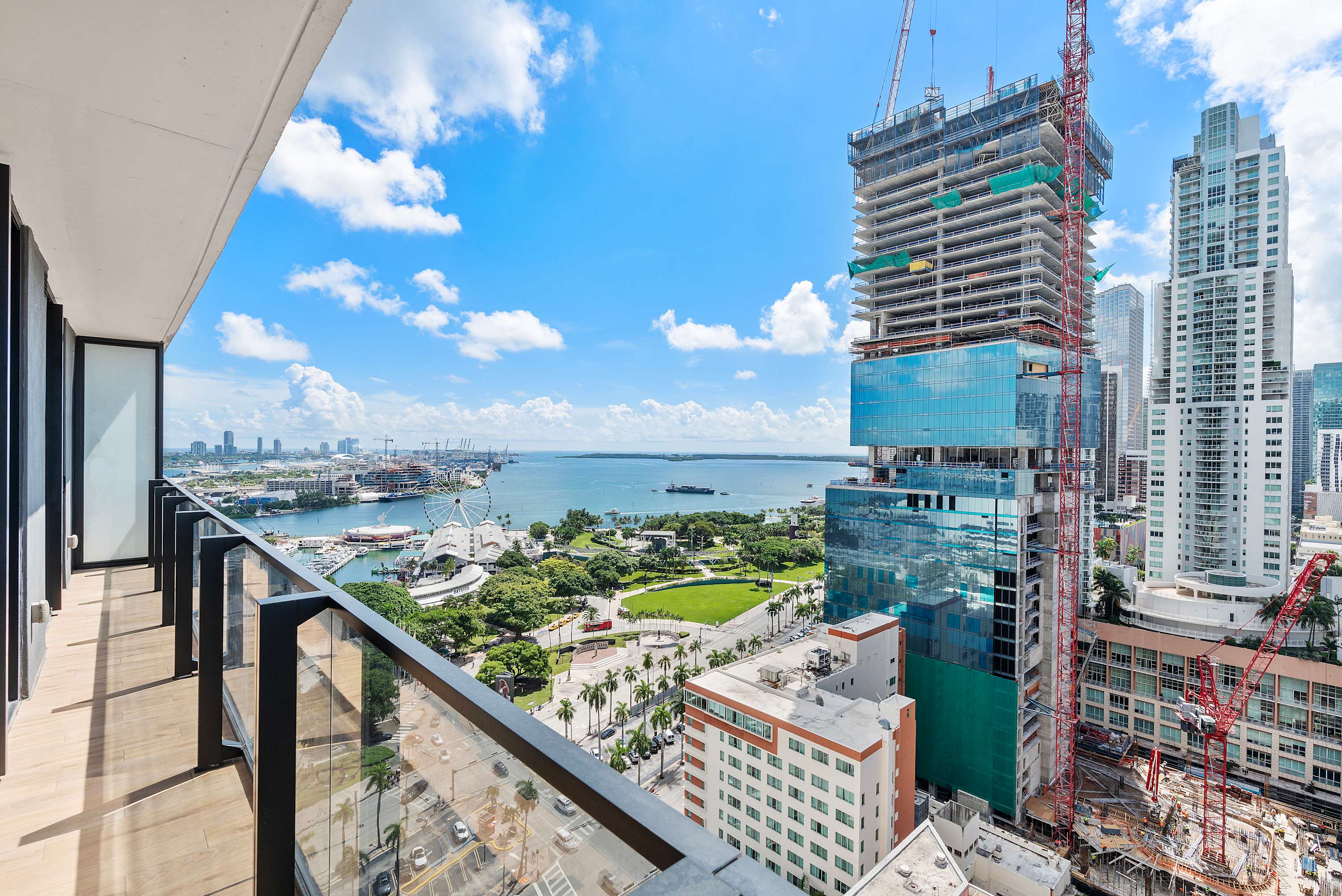 A high-angle view from a modern balcony with a glass railing overlooks a bustling city, a wide bay with boats, and a large Ferris wheel, with a prominent construction site in the foreground under a bright blue sky.