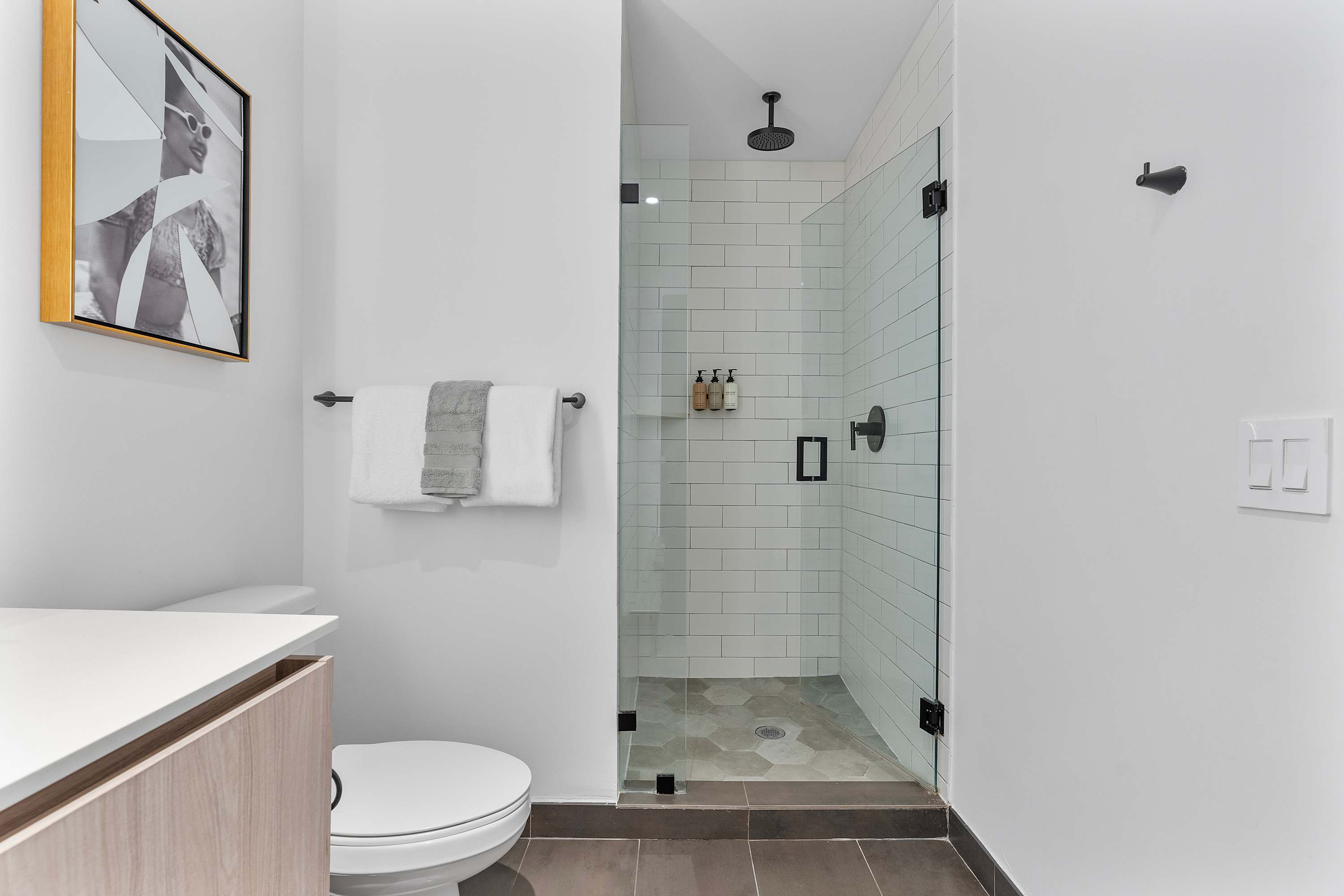 Modern walk-in shower with white subway tiles and a glass enclosure, featuring black fixtures including a rain showerhead. The shower floor has hexagonal gray tiles.