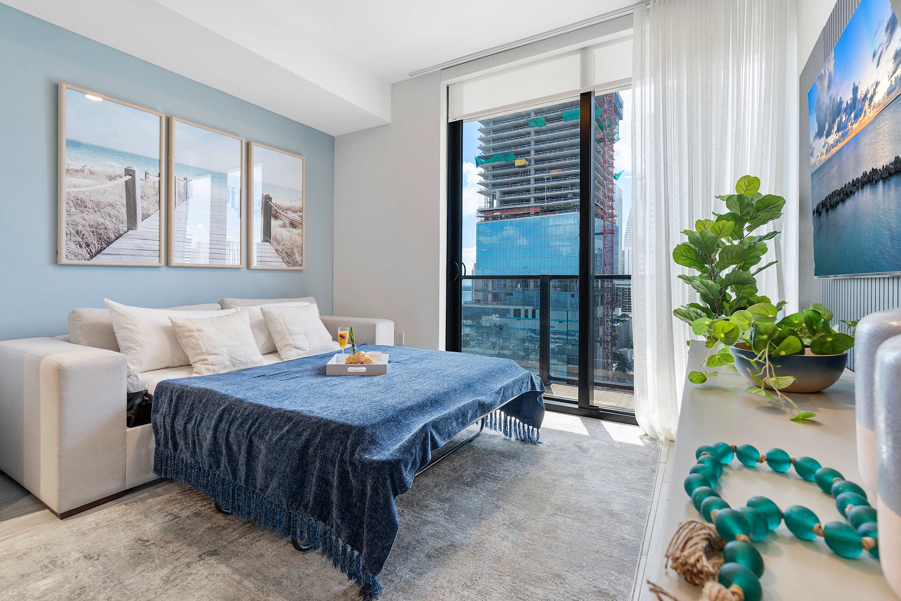 Living room converted into a sleeping area with a sofa bed made up with a blue blanket, light blue accent wall, and large windows offering city views, complemented by beach-themed artwork.