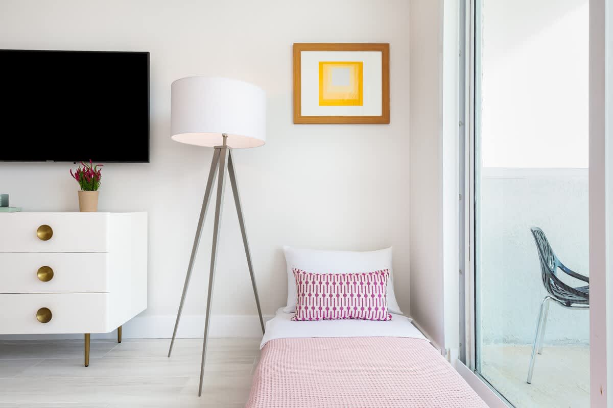 A versatile grey daybed, adorned with a pink blanket and patterned pillow, is positioned next to a large window, providing extra sleeping space in a bright, modern room. A tripod floor lamp and a white dresser with a flat-screen TV are also visible.