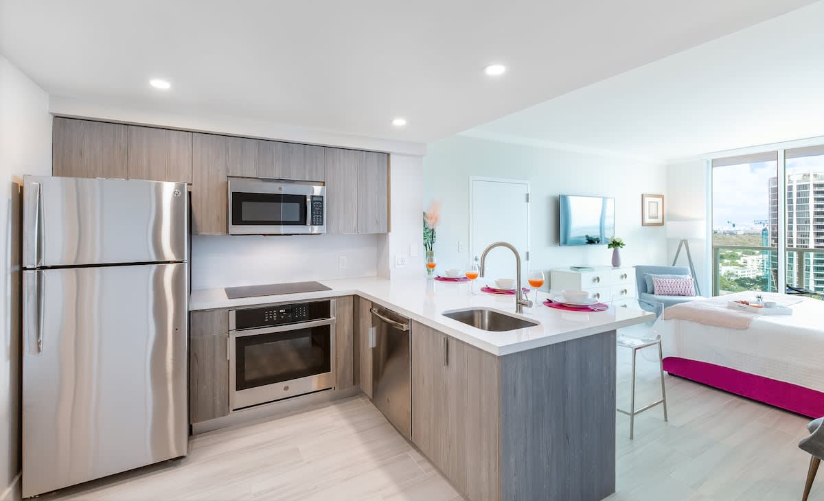 A sleek, modern kitchen features light wood and grey cabinetry, stainless steel appliances including a refrigerator, microwave, oven, and dishwasher, all centered around a white quartz island with an integrated sink. The open layout connects seamlessly to the rest of the studio.