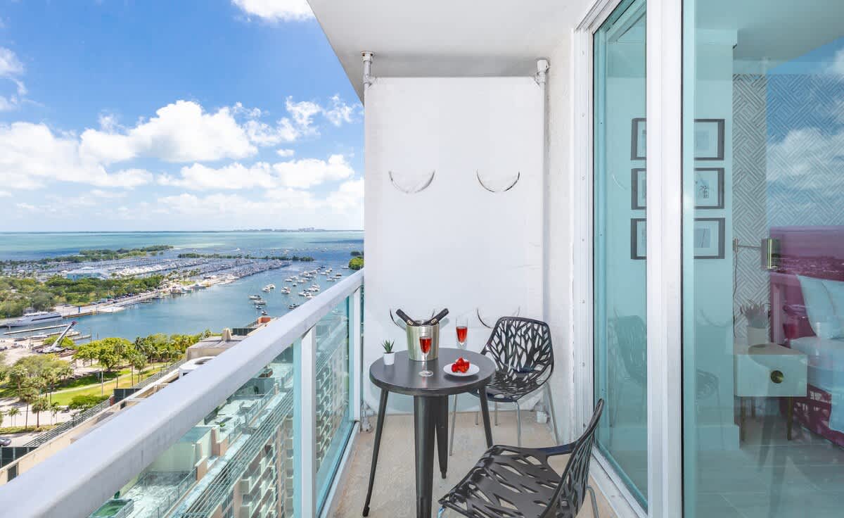 High-rise balcony with glass railings offering panoramic views of a bay filled with boats and a city skyline, furnished with a small black bistro table and two chairs.