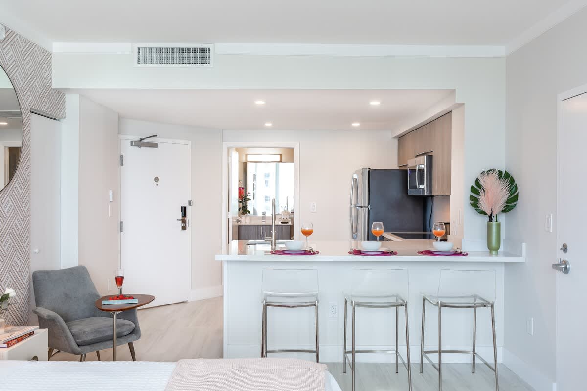 A wide-angle view from the bed showcases the open-concept studio, featuring a modern kitchen with a breakfast bar and clear bar stools, leading to the entrance door. A grey armchair and a round mirror are visible on the left, adding to the contemporary decor.