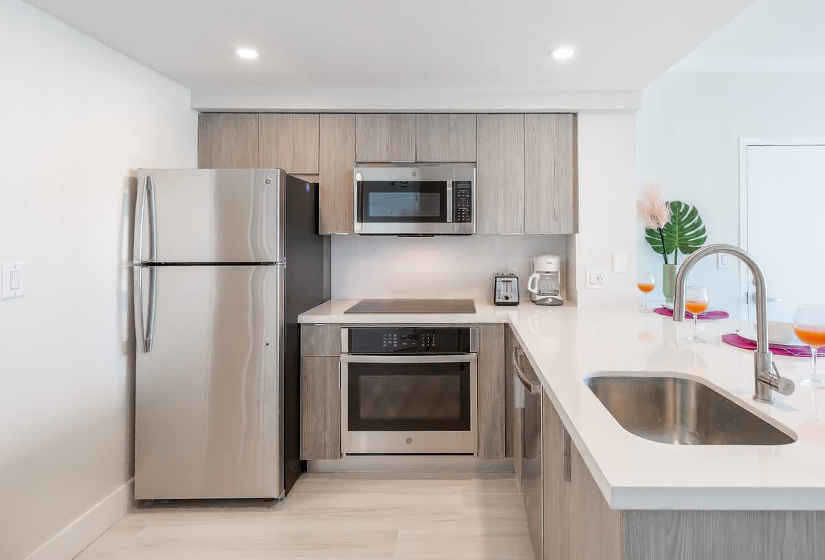 Modern kitchen with light wood-grain cabinetry, stainless steel appliances including a refrigerator, microwave, oven, and electric cooktop, and a white quartz countertop with an undermount sink.