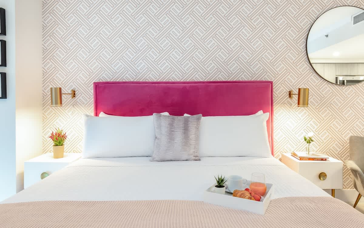 A cozy bedroom features a white bed with a vibrant fuchsia velvet headboard, flanked by white nightstands with gold accents and brass wall sconces. A geometric patterned wallpaper adds texture behind the bed, creating a stylish and inviting space.