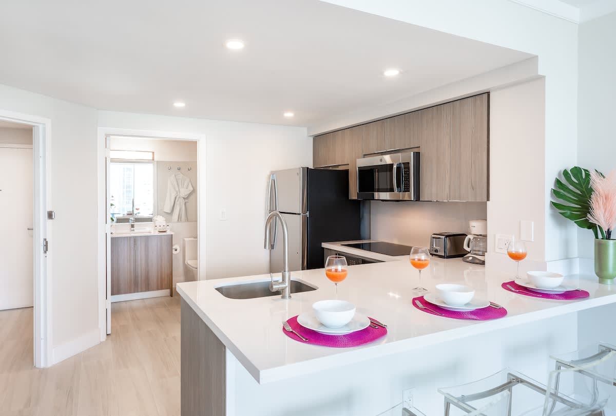 A modern kitchen features light wood cabinetry, stainless steel appliances, and a white quartz countertop with a breakfast bar set for dining. Beyond the kitchen, a bright bathroom with a vanity and robes is visible through an open doorway.