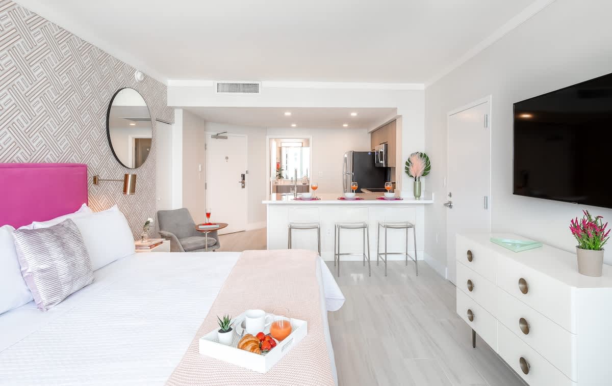 A modern studio apartment combines a bedroom area with a white bed and fuchsia headboard, a white dresser with a flat-screen TV, and an open-concept kitchen with a breakfast bar and clear bar stools. Light wood-look flooring unifies the bright, contemporary space.