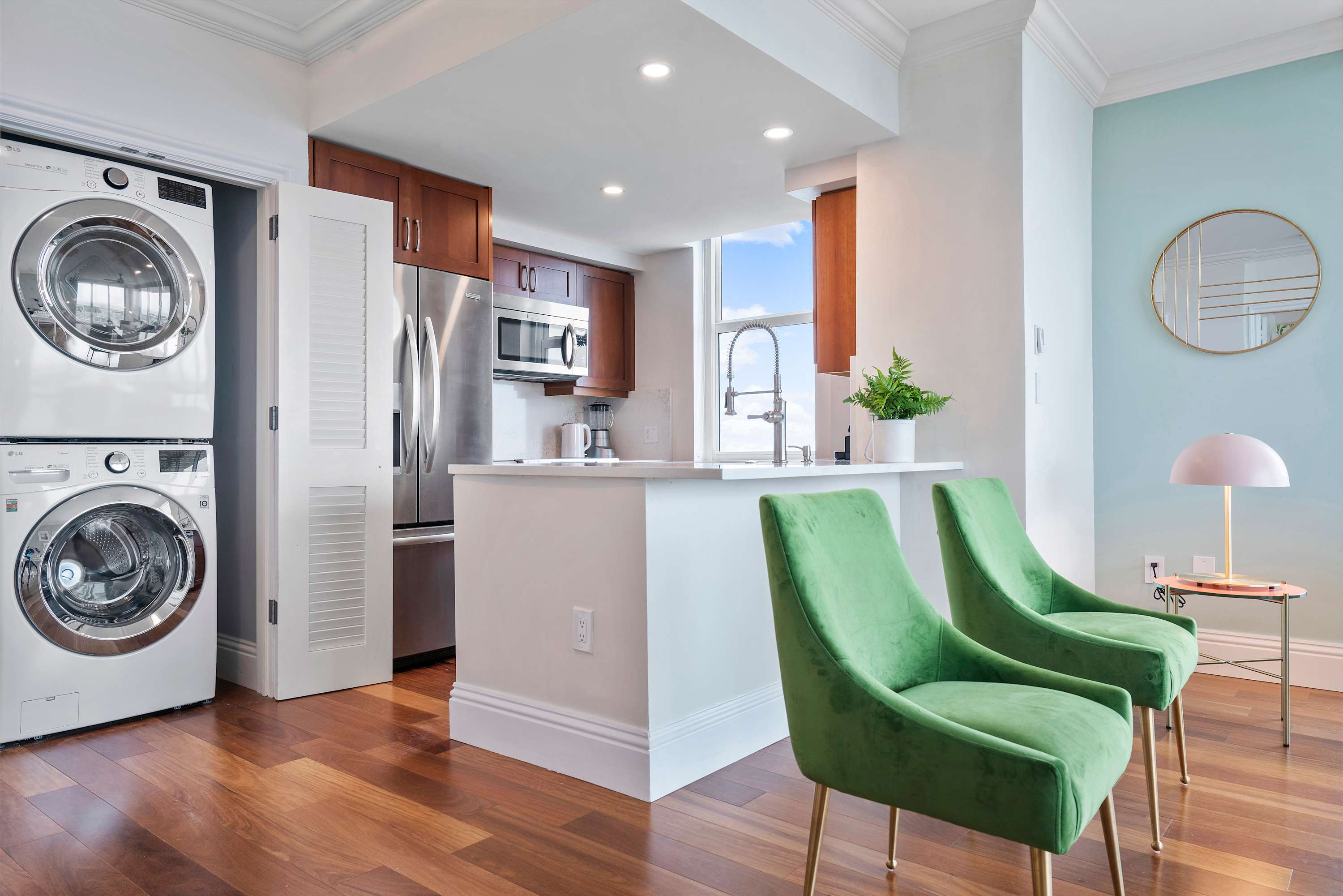 An open-concept kitchen and living area with hardwood floors, featuring a stacked washer and dryer unit in a closet. The kitchen has stainless steel appliances and a breakfast bar with two green velvet chairs.