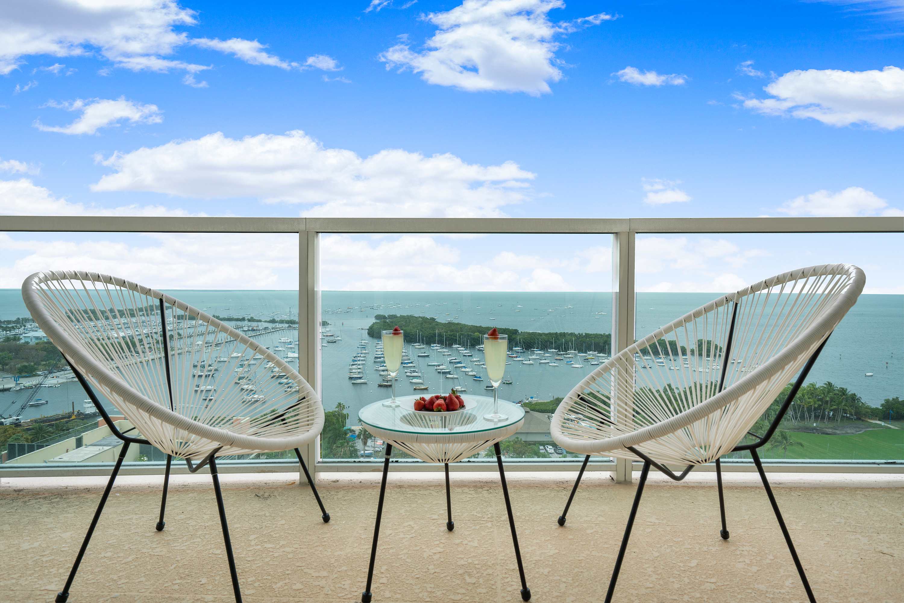 A close-up view of a private balcony featuring two white woven chairs and a small glass table, offering a serene vista of the bay dotted with numerous boats. Two glasses of champagne and strawberries are on the table.