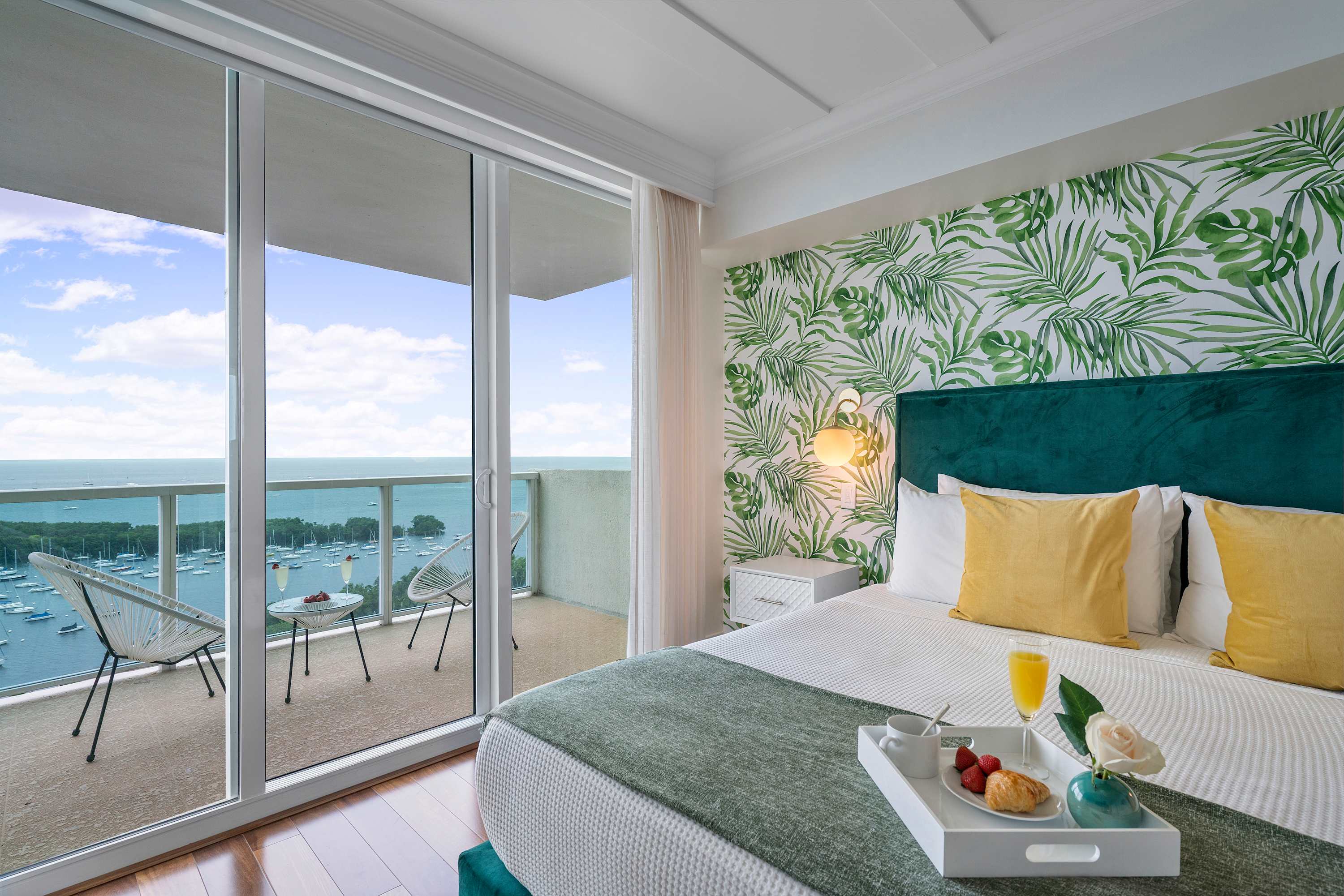 A stylish bedroom with a teal upholstered bed, a tropical leaf-patterned accent wall, and hardwood floors. Sliding glass doors lead to a private balcony with views of the bay and boats.