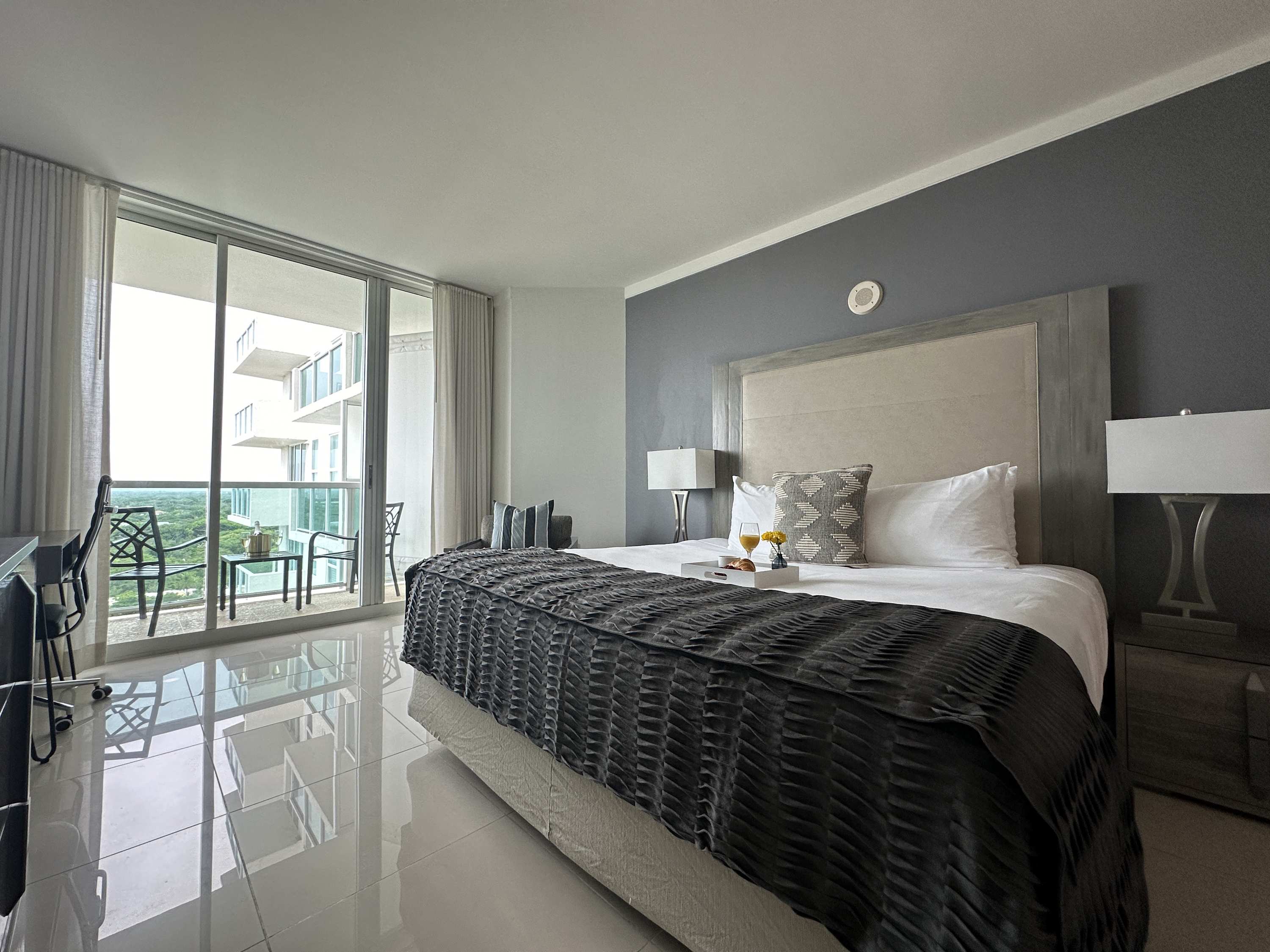 A spacious studio apartment with a king-sized bed, a small desk and chair, and large sliding glass doors opening to a balcony. The room features light tile flooring and a modern aesthetic.