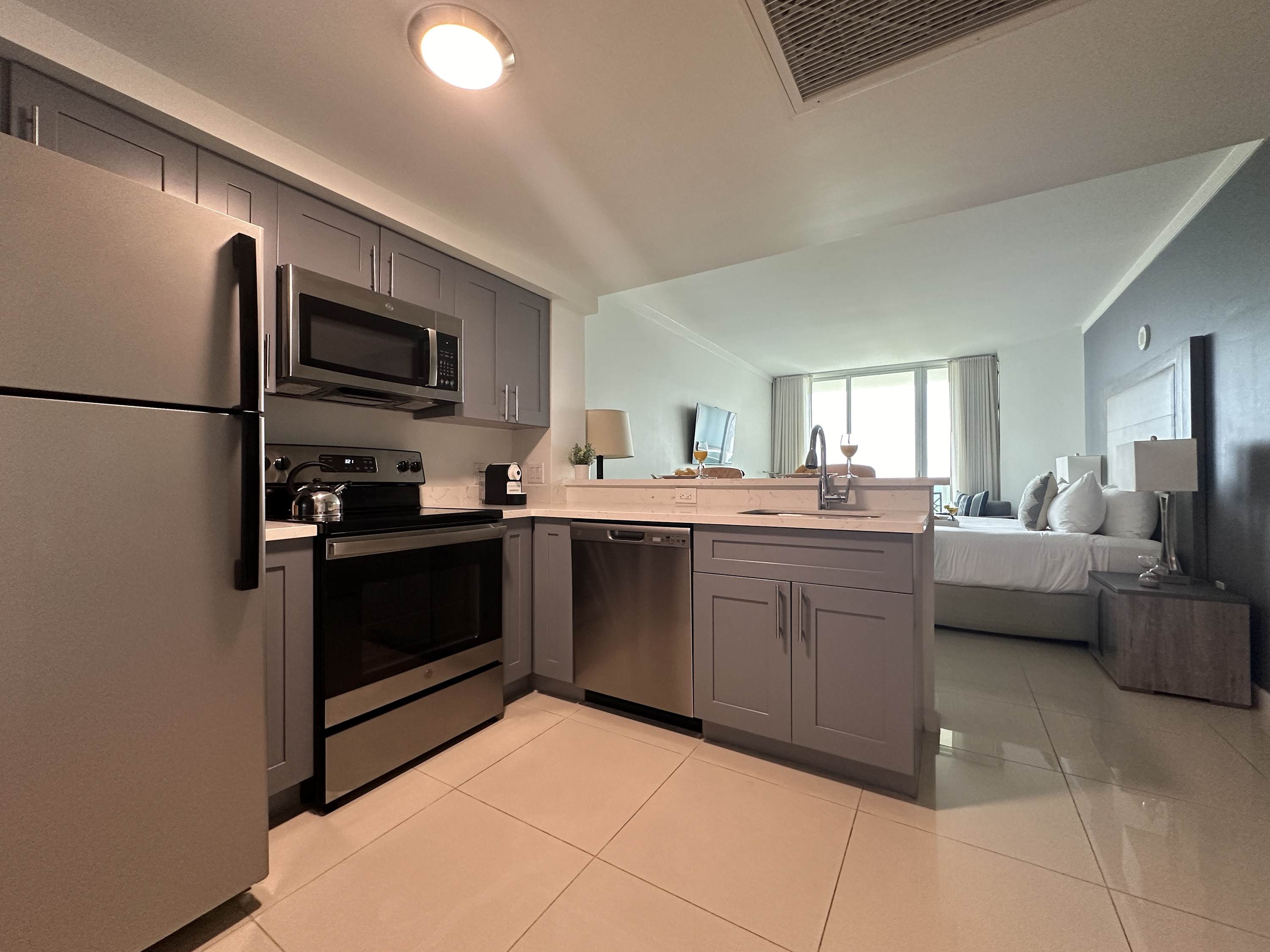 A well-equipped modern kitchen with gray shaker-style cabinets, stainless steel appliances including a full-sized refrigerator, stove, and microwave, and a breakfast bar, seamlessly connected to the studio living space.