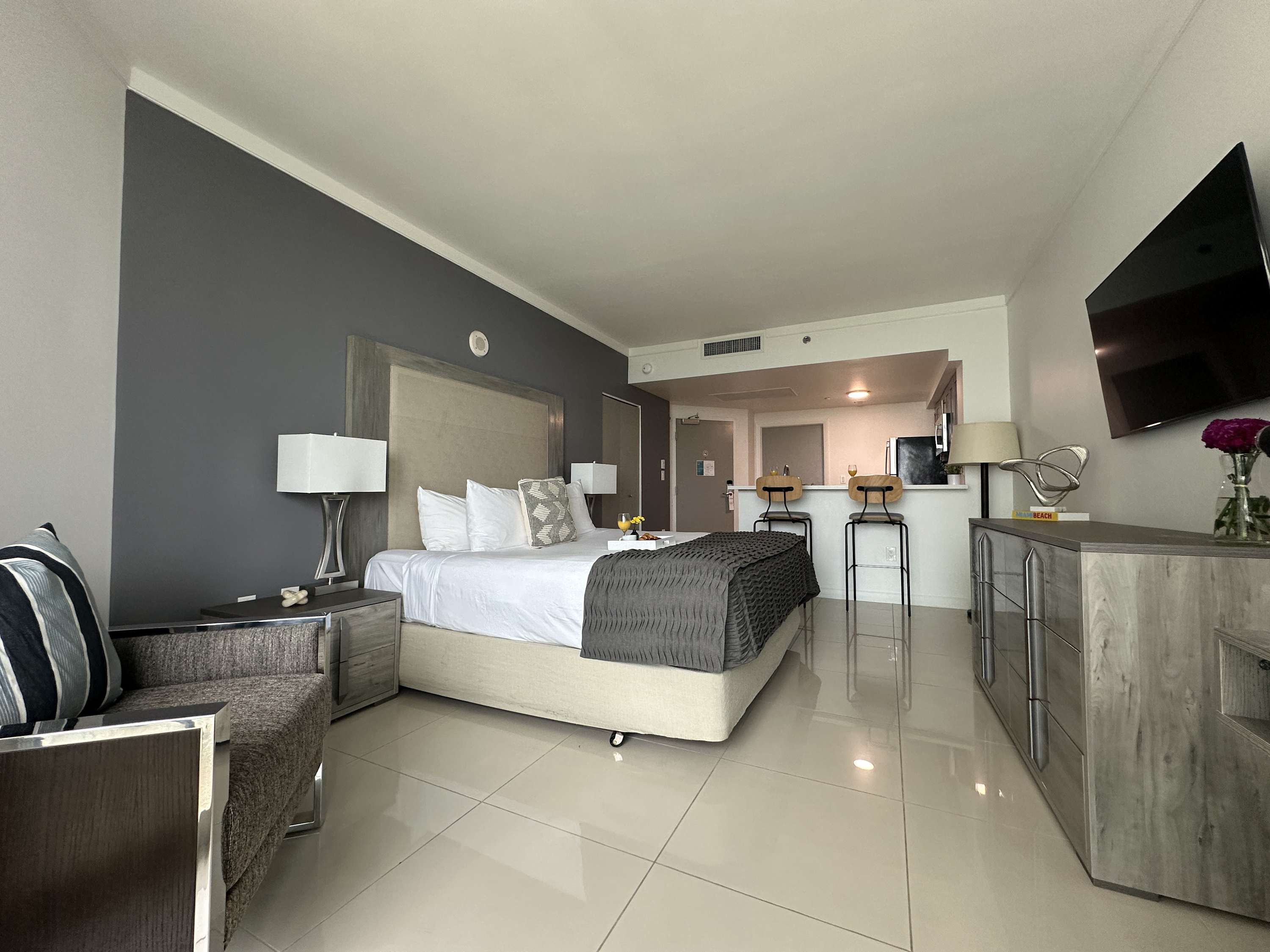 A wide shot of the studio apartment showcasing a king-sized bed, a small gray armchair, and an open-concept kitchen area with a breakfast bar, all set on light tile flooring.