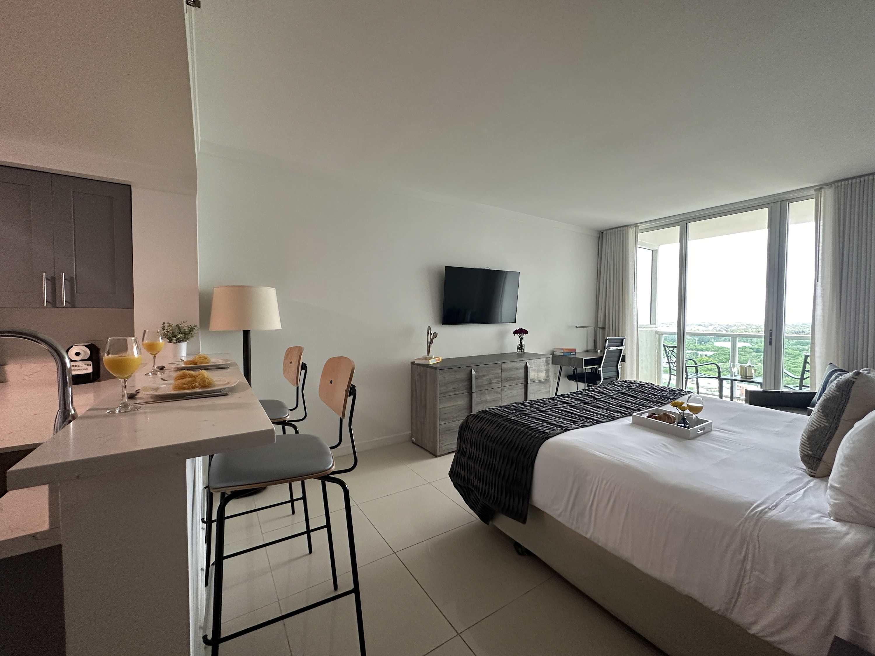 A bright studio apartment with a king-sized bed, a small breakfast bar with two stools, and a flat-screen TV. Large sliding glass doors lead to a balcony with an outdoor seating area, offering a scenic view.