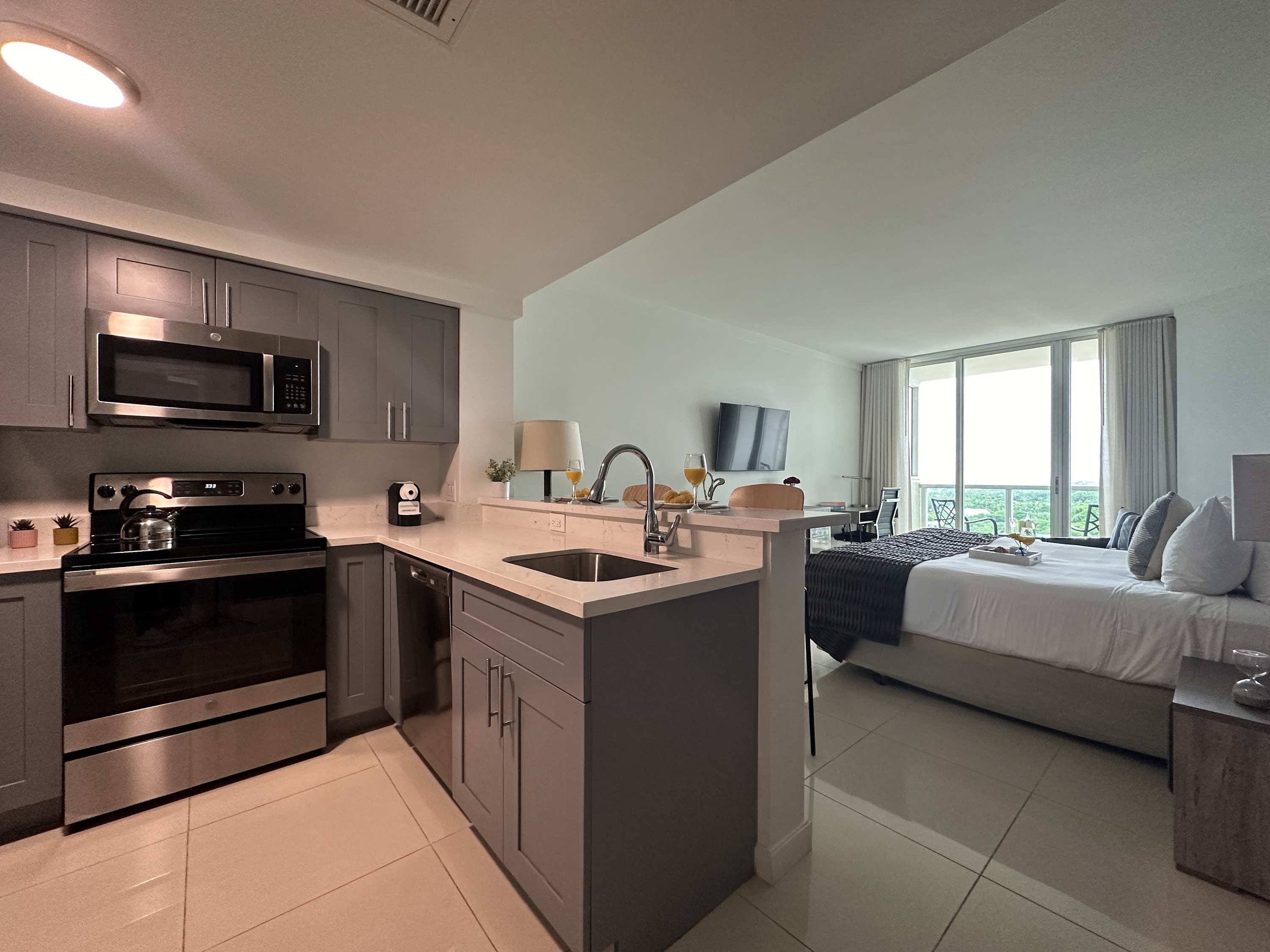 A modern kitchen featuring gray shaker-style cabinets, stainless steel appliances including a microwave, stove, and refrigerator, with light tile flooring and an open view into the adjacent studio living space.