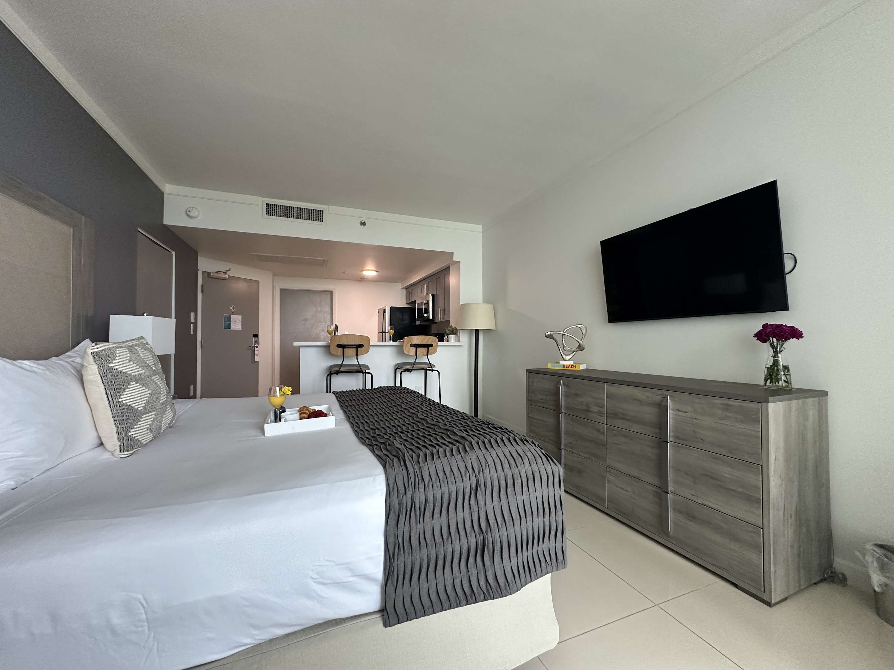 A modern studio apartment featuring a king-sized bed with a gray textured throw, a large flat-screen TV mounted above a gray dresser, and a glimpse of the kitchen/bar area in the background.