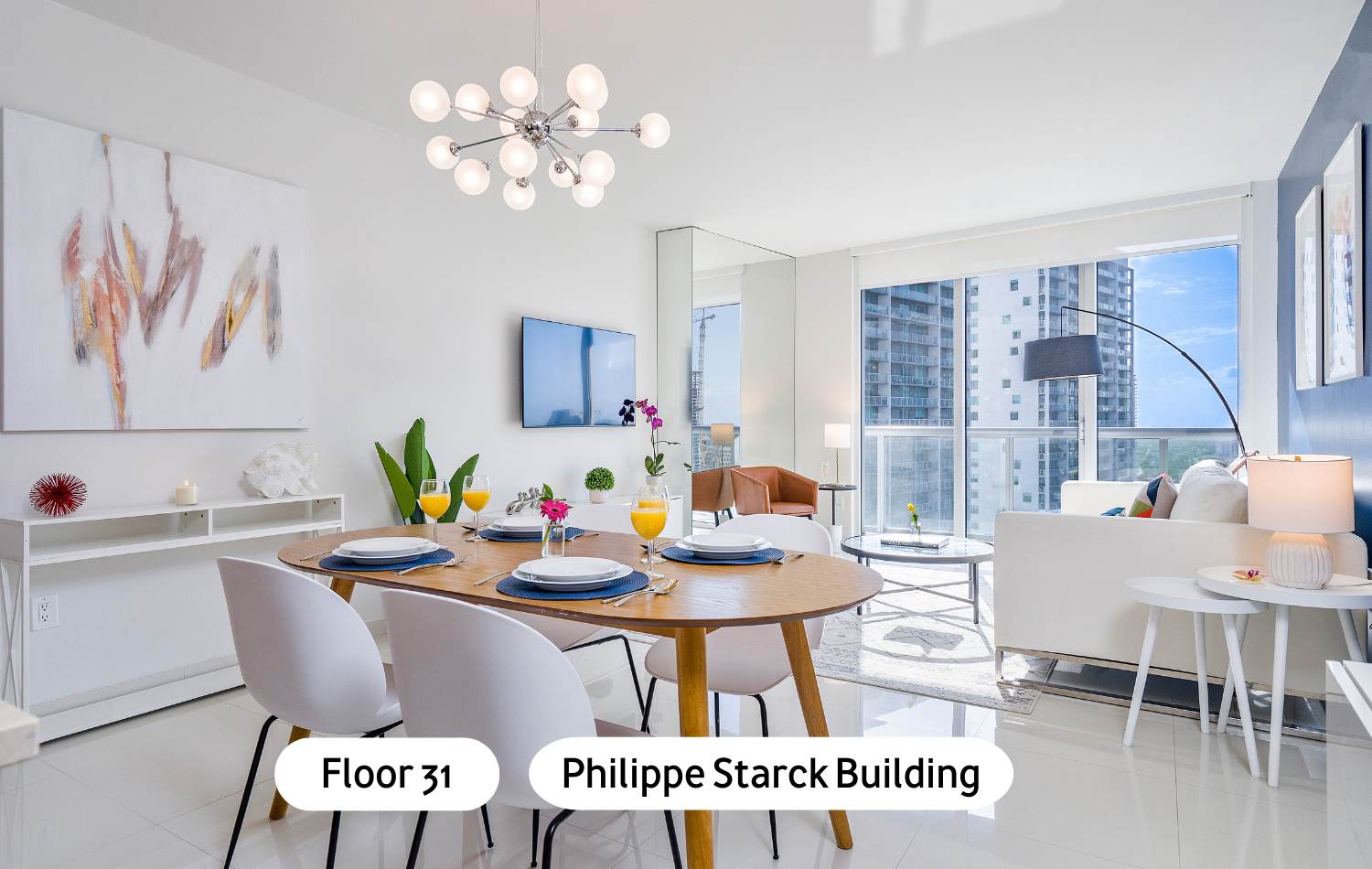 Modern open-concept dining area featuring an oval wooden table with white chairs, a Sputnik-style chandelier, and an abstract painting, flowing into a living space with city views through large windows.