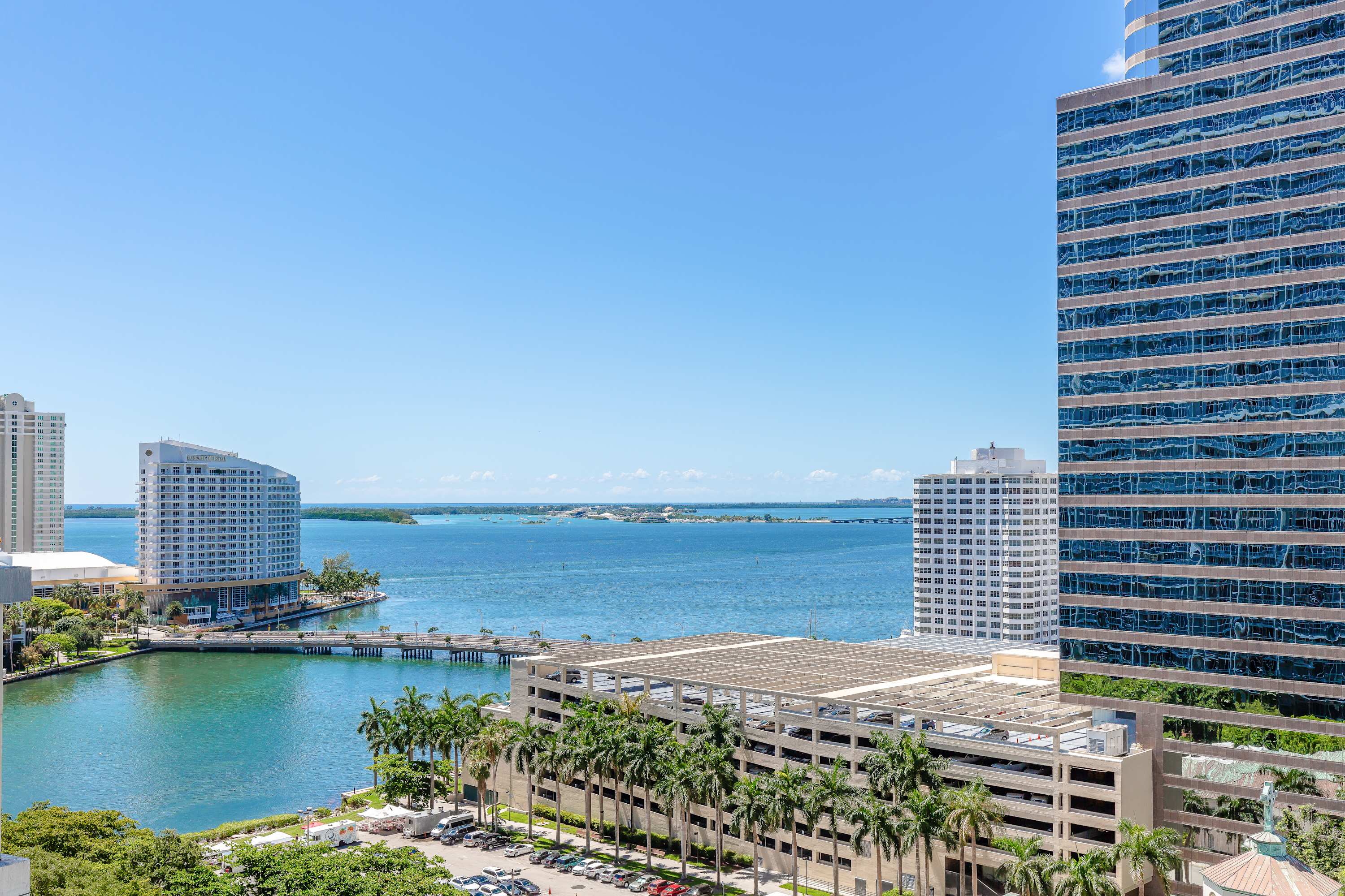 Expansive daytime view of a city waterfront, featuring a bay with a bridge, surrounding high-rise buildings, and a parking garage. Palm trees line the water's edge under a clear blue sky.