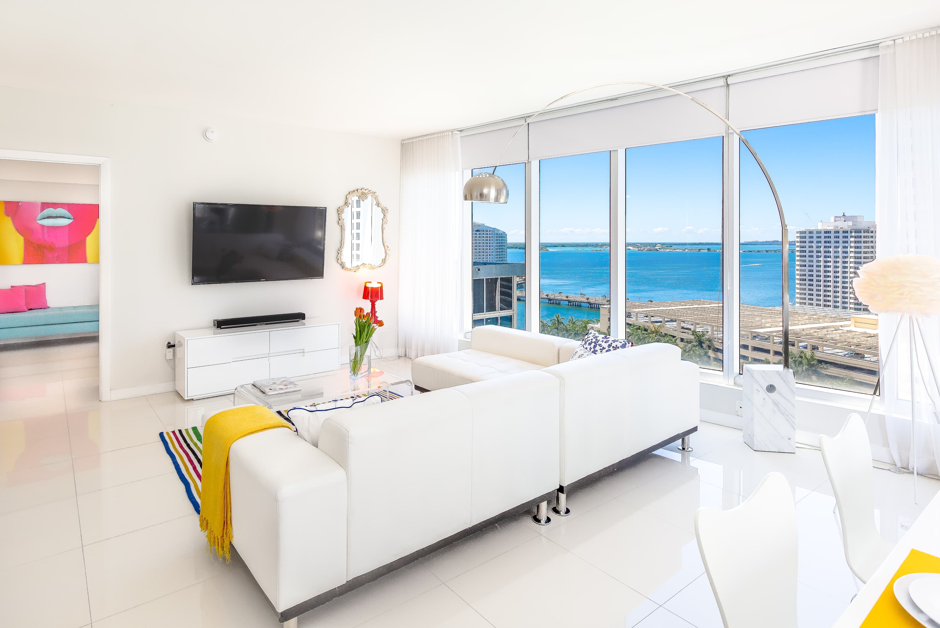 Spacious living room with a white sectional sofa, wall-mounted TV, and floor-to-ceiling windows showcasing expansive ocean and city views. A colorful throw blanket and a unique arc lamp add modern touches to the bright interior.