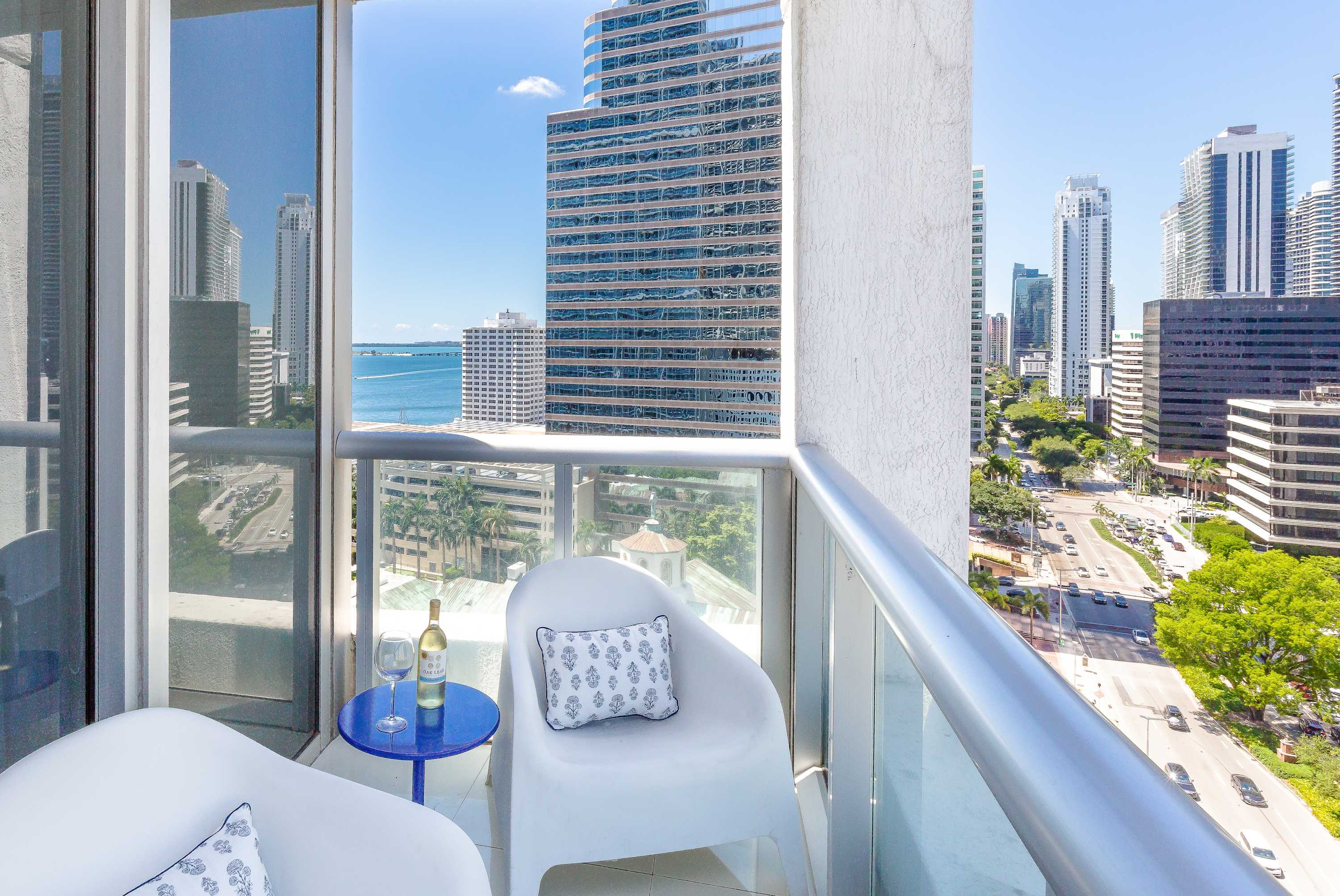 Private balcony offering panoramic city and waterfront views, furnished with two modern white chairs and a small blue side table. A bottle of wine and glasses are set on the table.