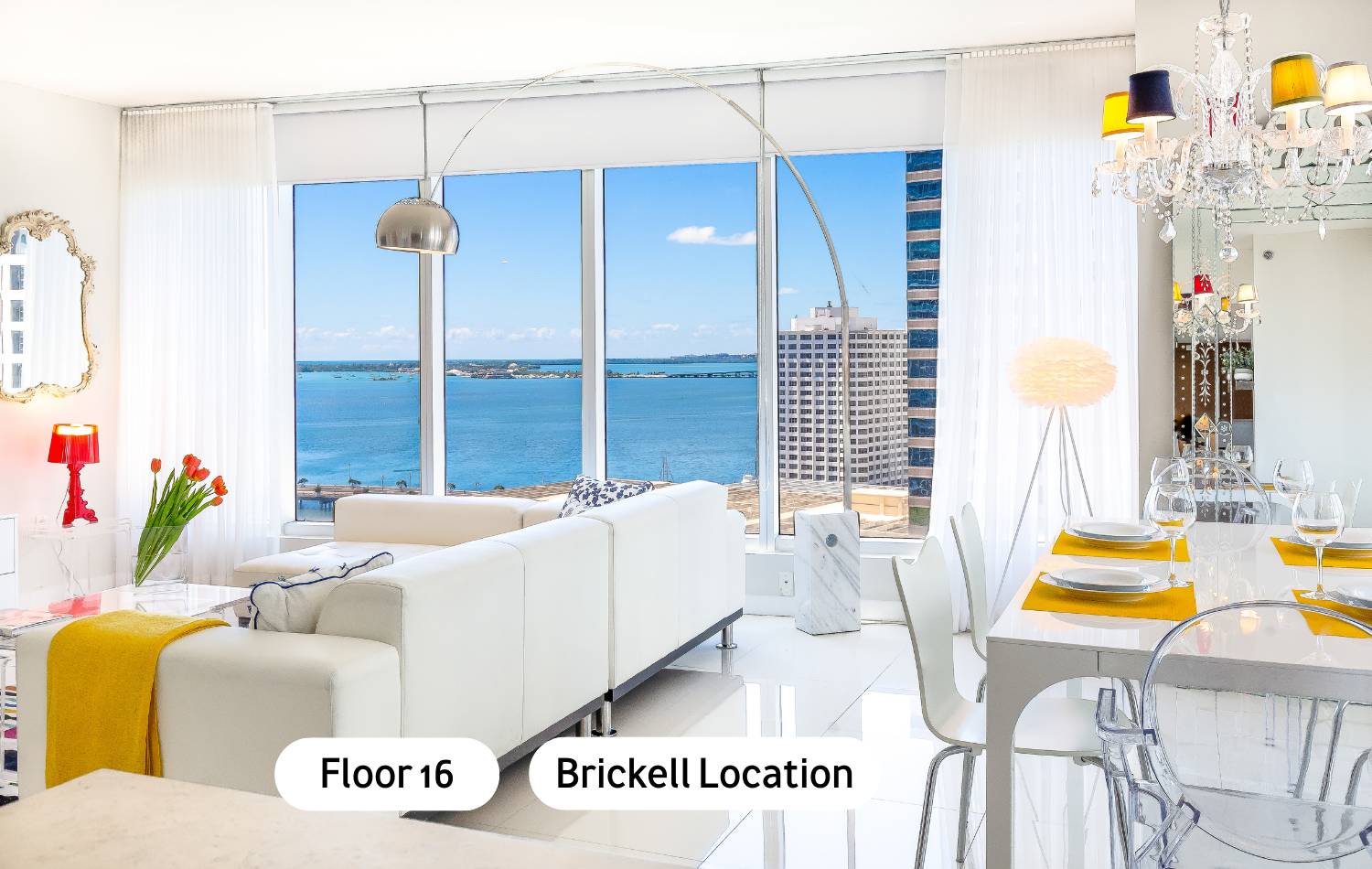 Modern open-concept living and dining area with a white leather sofa, white dining table, and floor-to-ceiling windows offering panoramic ocean and city views. A large arc lamp and a colorful chandelier add character to the bright space.
