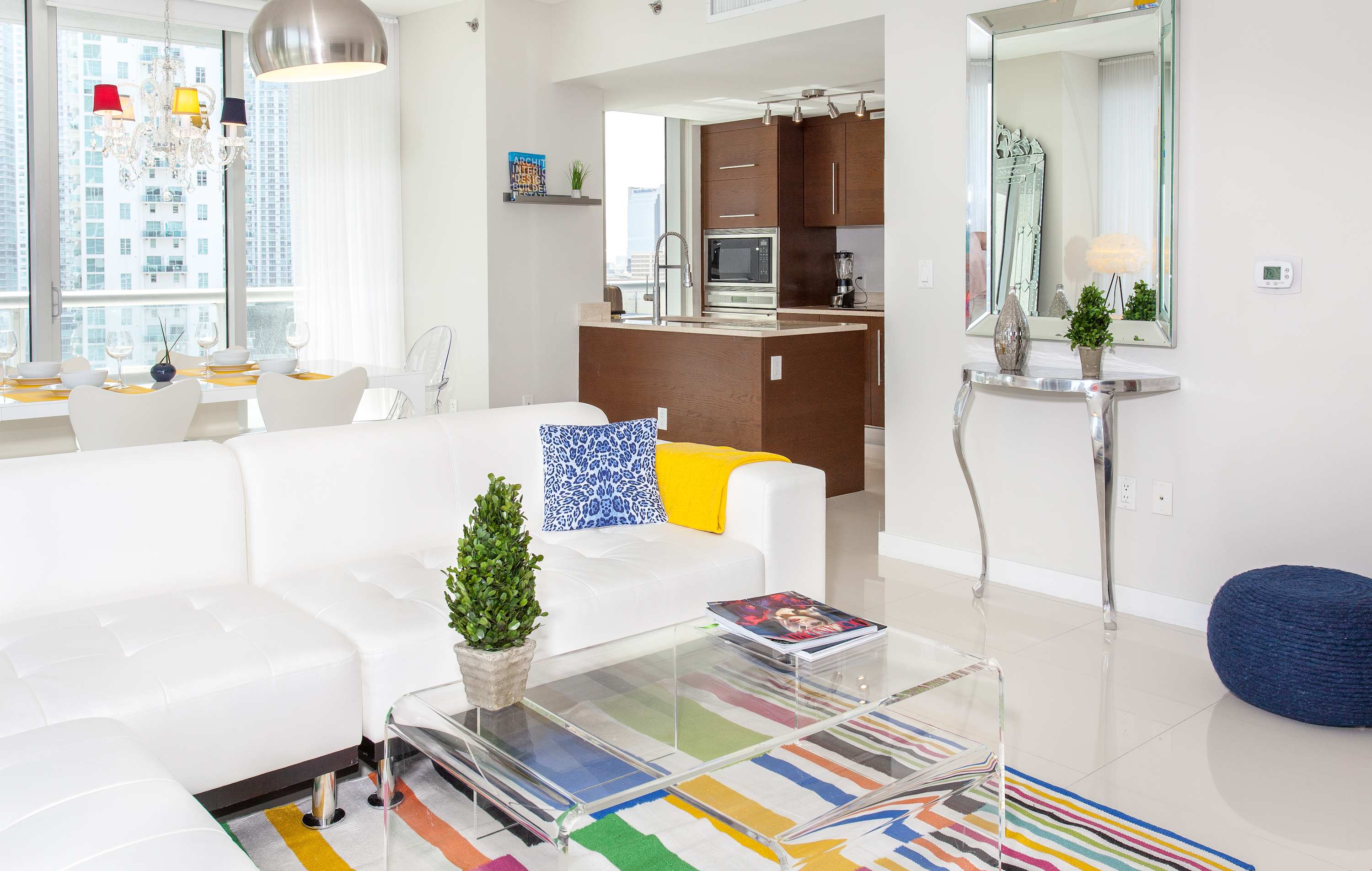 Stylish living room with a white sectional sofa, a clear coffee table, and a vibrant rug, offering a glimpse of the modern kitchen island and dining area. A tall, ornate silver mirror and a console table add decorative accents to the bright, open space.