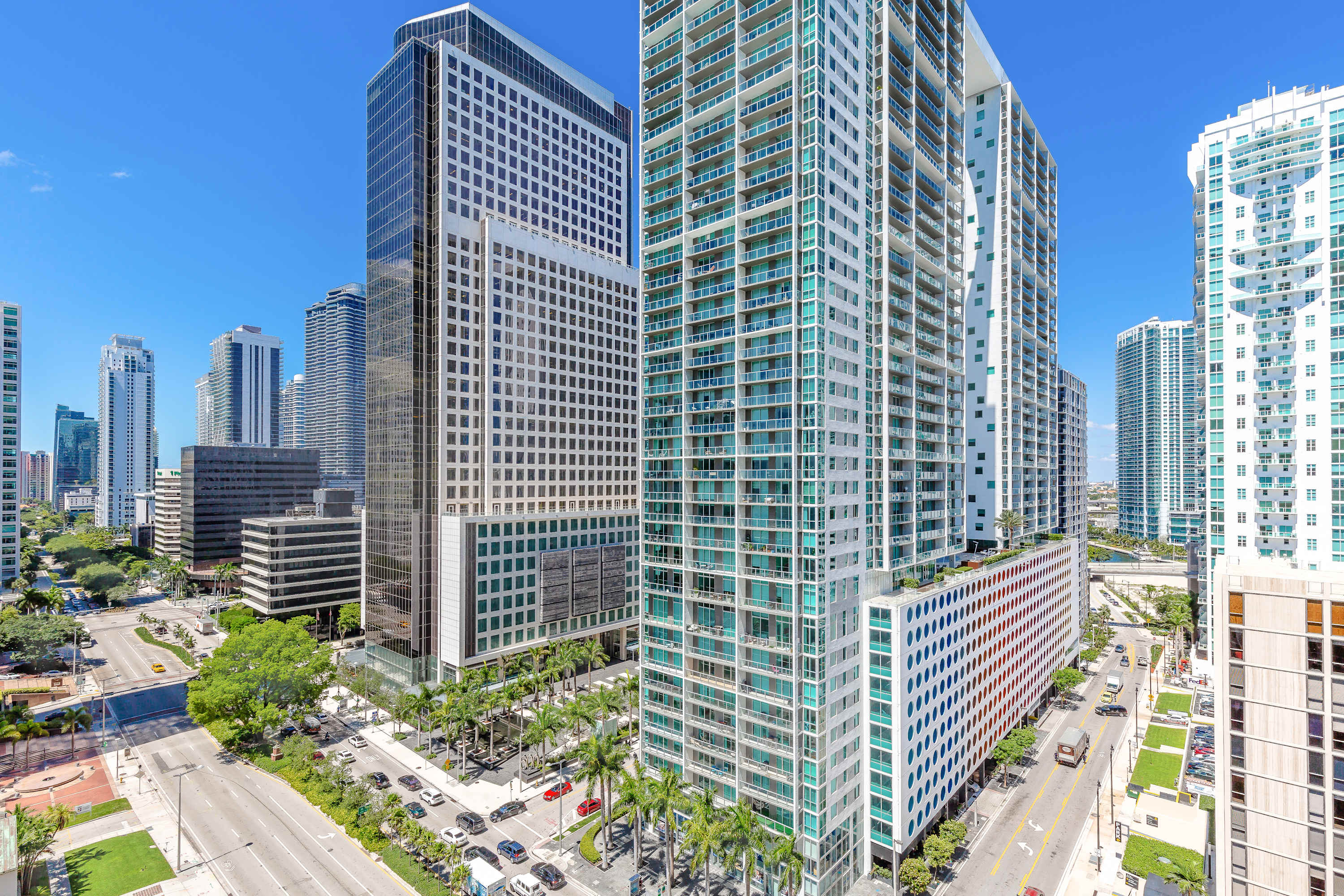 A panoramic high-floor view of a vibrant city, showcasing numerous modern skyscrapers, busy streets with cars, and lush palm trees under a clear blue sky.
