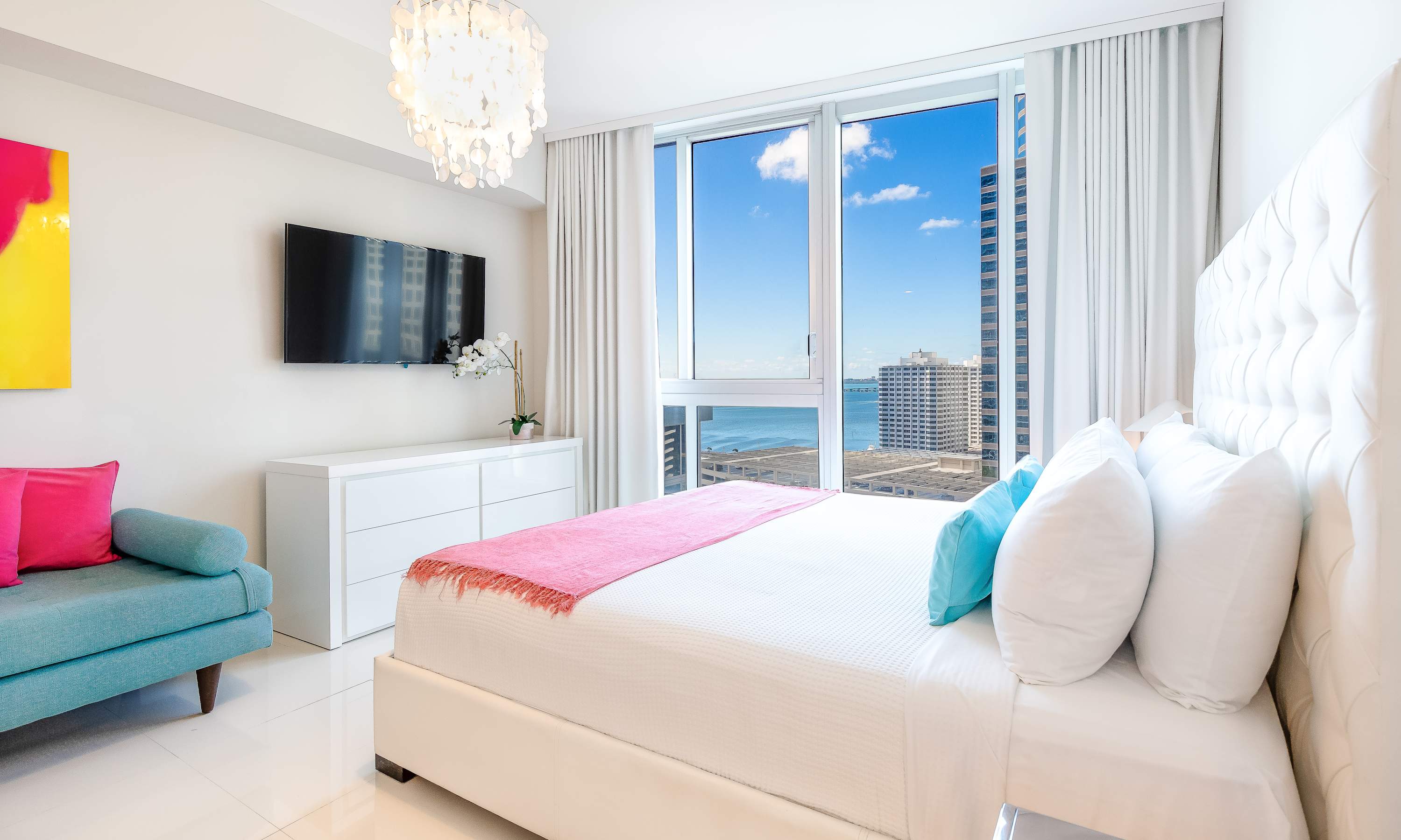Elegant bedroom with a white tufted headboard bed, pink throw blanket, and floor-to-ceiling windows offering city and water views. A wall-mounted TV, white dresser, and a teal accent chair complete the modern decor.
