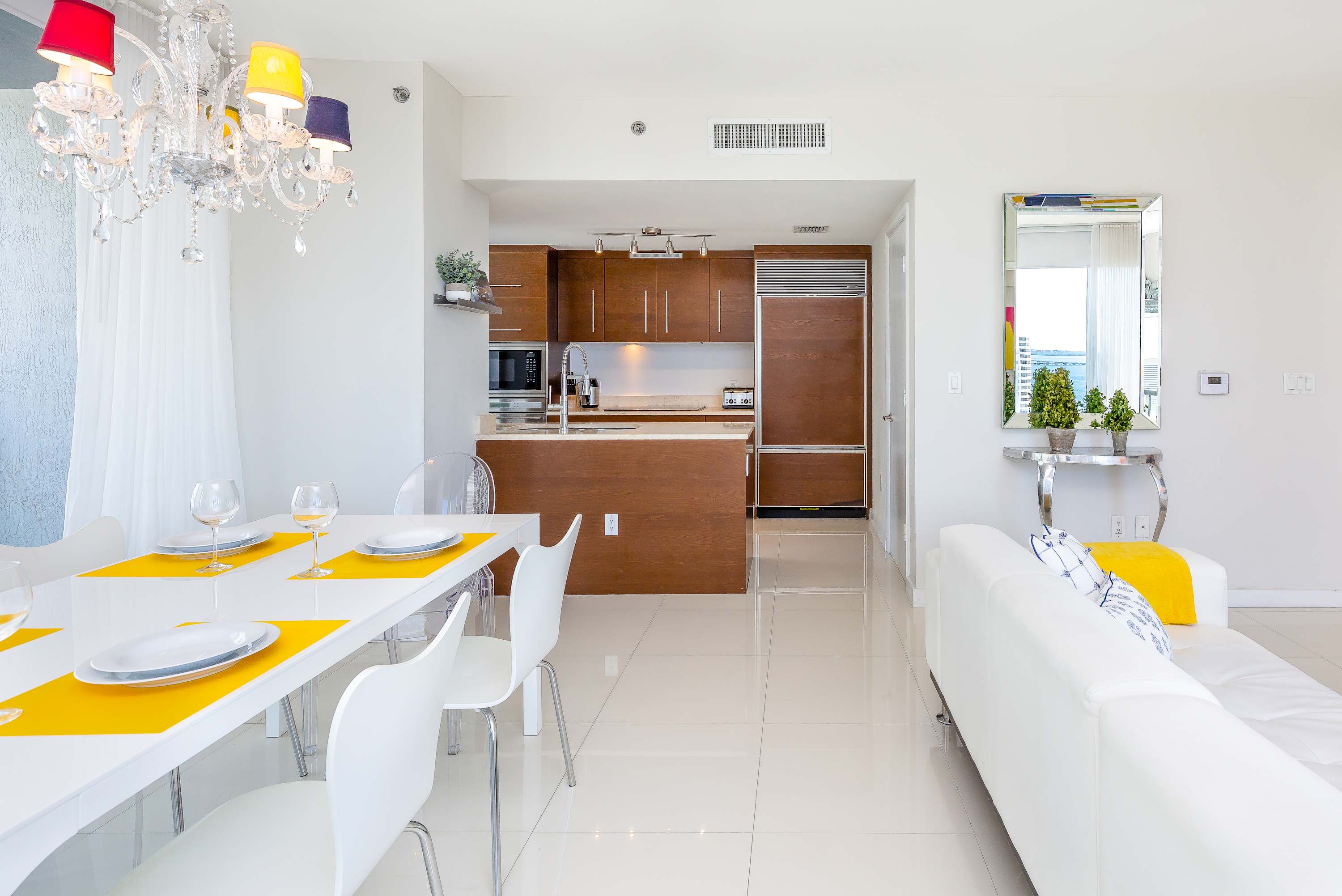 Bright open-concept dining area with a white table set for four, leading into a modern kitchen with dark wood cabinetry and stainless steel appliances. A large mirror and a colorful chandelier are prominent design elements.