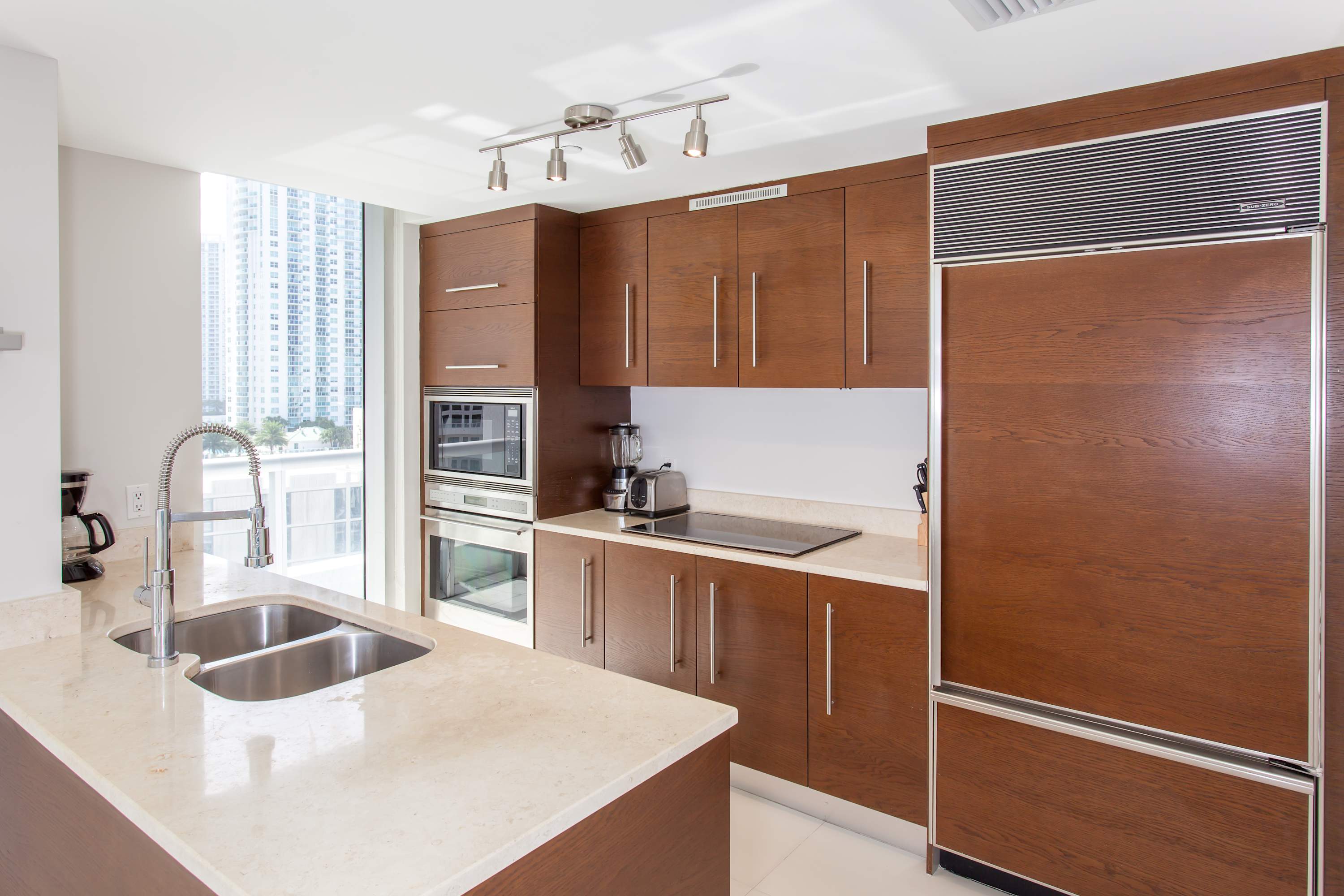 Contemporary kitchen featuring rich wooden cabinetry, light-colored countertops, and integrated stainless steel appliances, including a large built-in refrigerator. A track light fixture illuminates the workspace, and a window provides natural light and a city view.