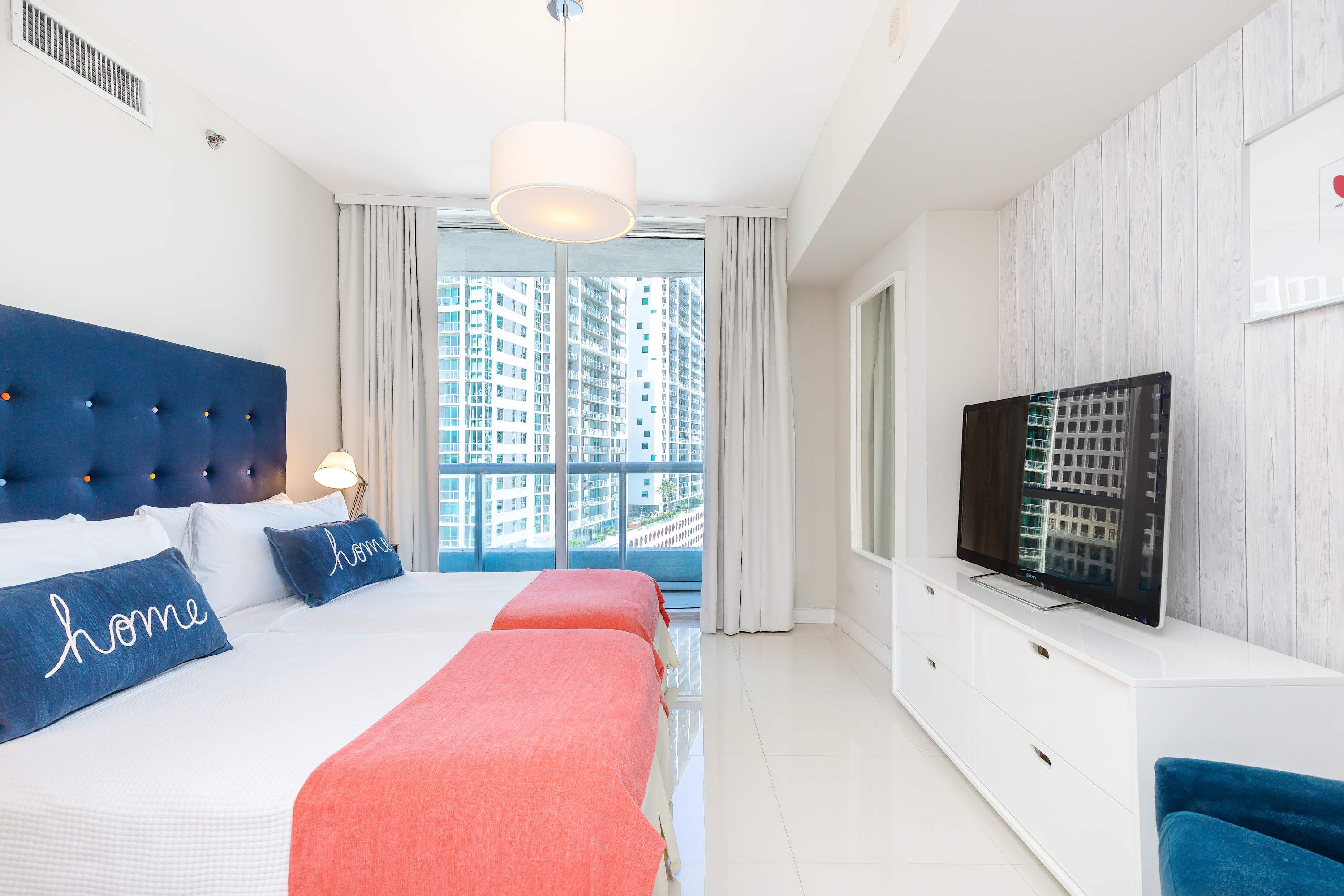 Bright bedroom with two single beds featuring navy blue headboards and coral throws, positioned against a large window offering city views. A white dresser with a flat-screen TV sits opposite the beds, and a modern pendant light illuminates the room.
