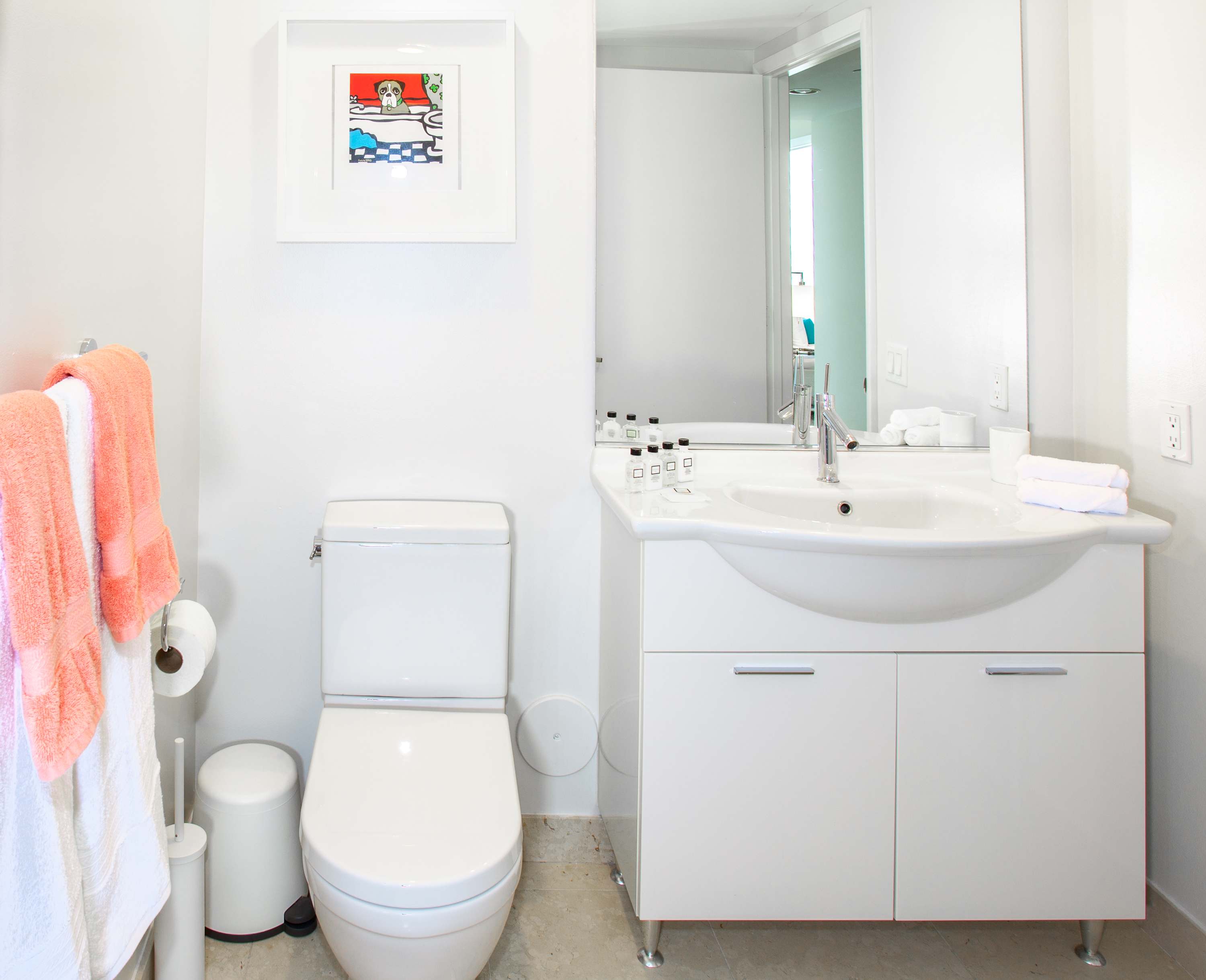 Compact modern bathroom with a white toilet and a sleek white vanity with an integrated sink and chrome faucet. A framed artwork hangs above the toilet, and orange and white towels are on a rack.